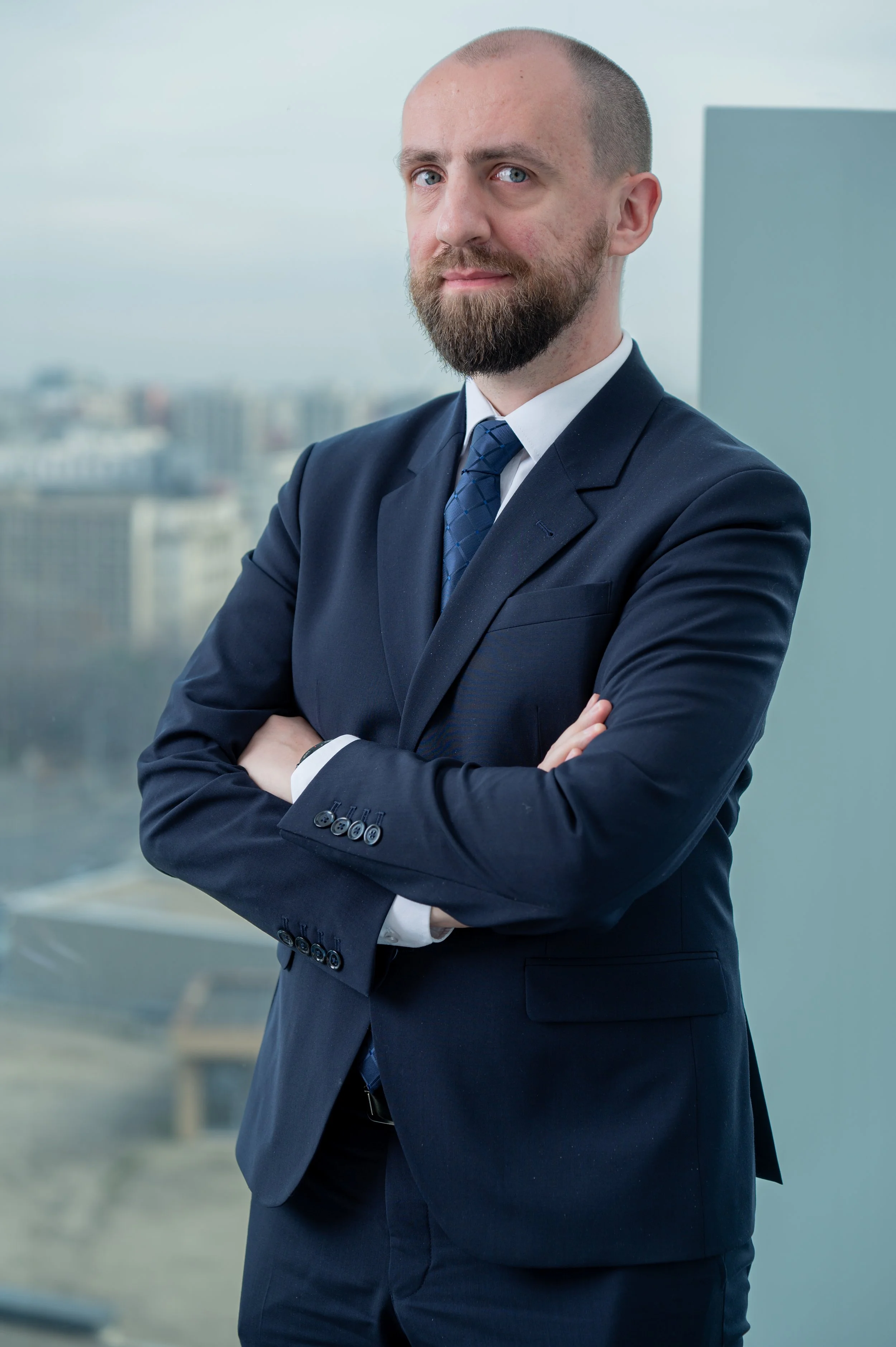 A man wearing a dark blue suit with crossed arms, standing in front of a blurred cityscape background with high-rise buildings, on a cloudy day.