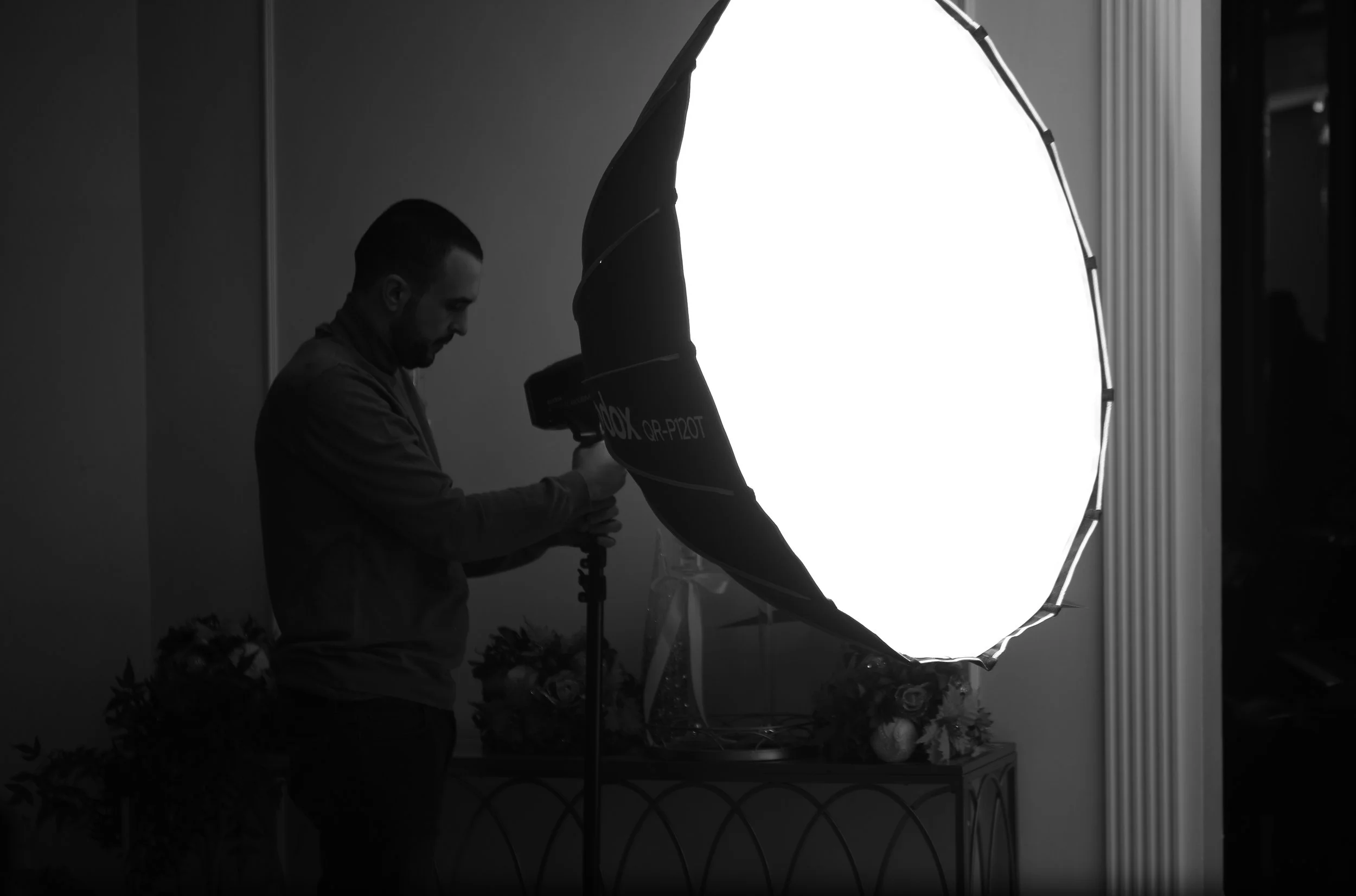 A person working with photographic equipment in a studio with a large softbox light. The scene is in black and white.