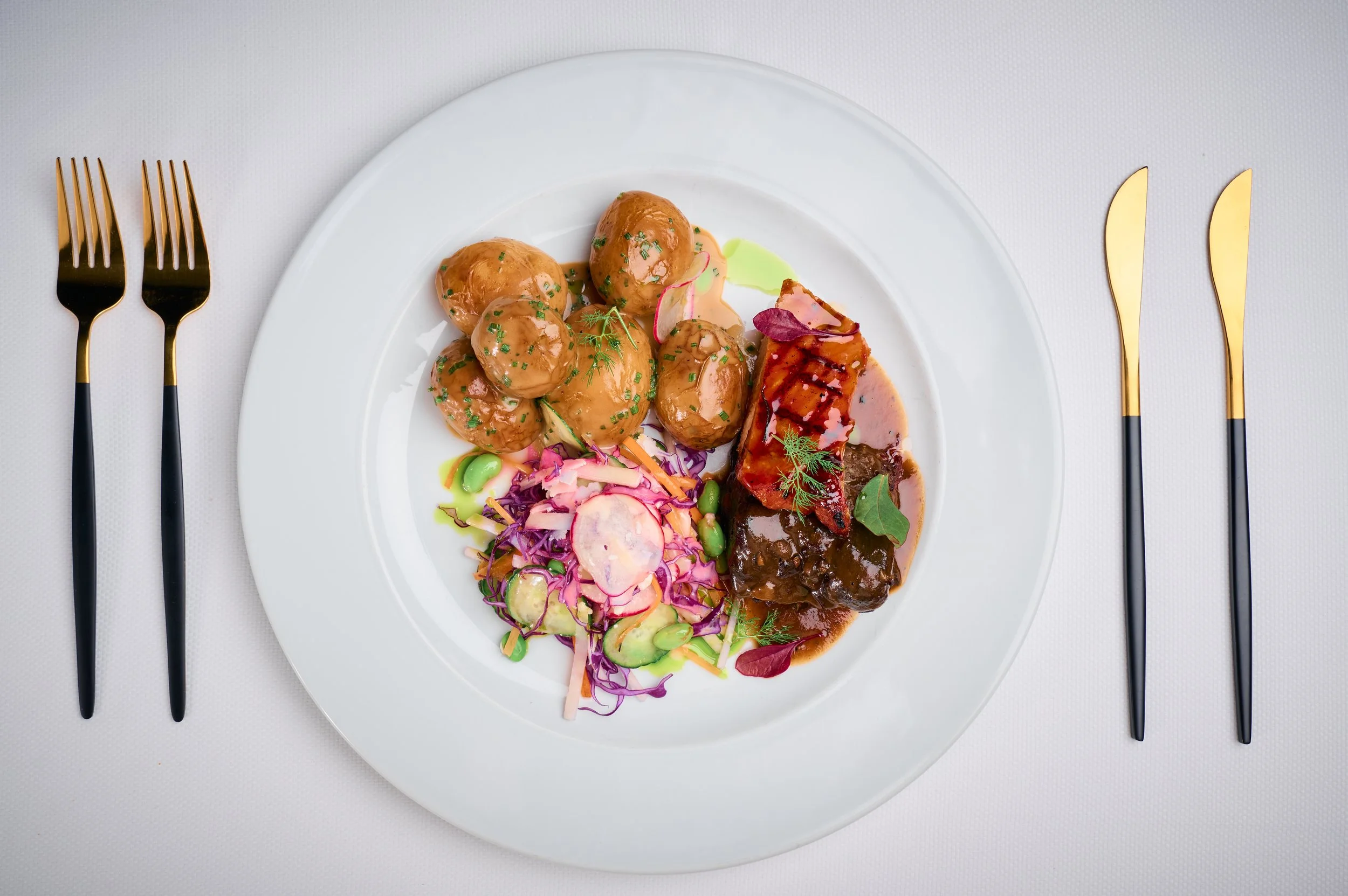 A white plate with roasted potatoes, glazed meat, and assorted vegetables, garnished with herbs, all set on a white tablecloth with black and gold cutlery on either side.