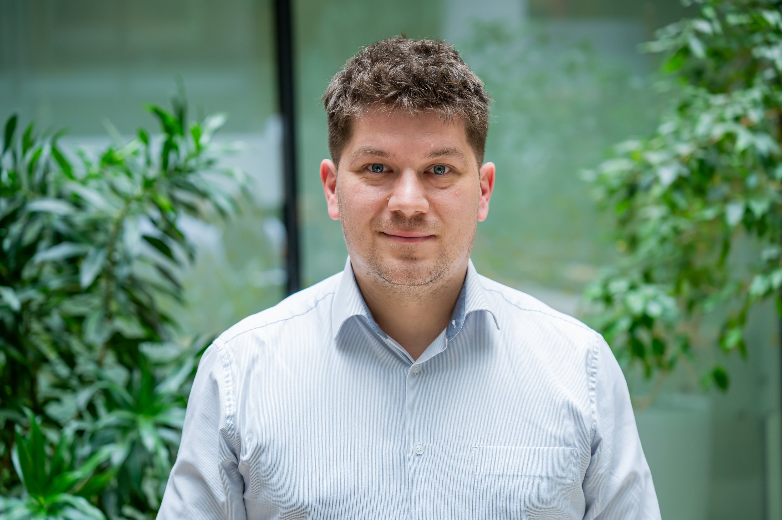 A man with short, brown hair and blue eyes is standing indoors, surrounded by green plants, wearing a light blue button-up shirt.