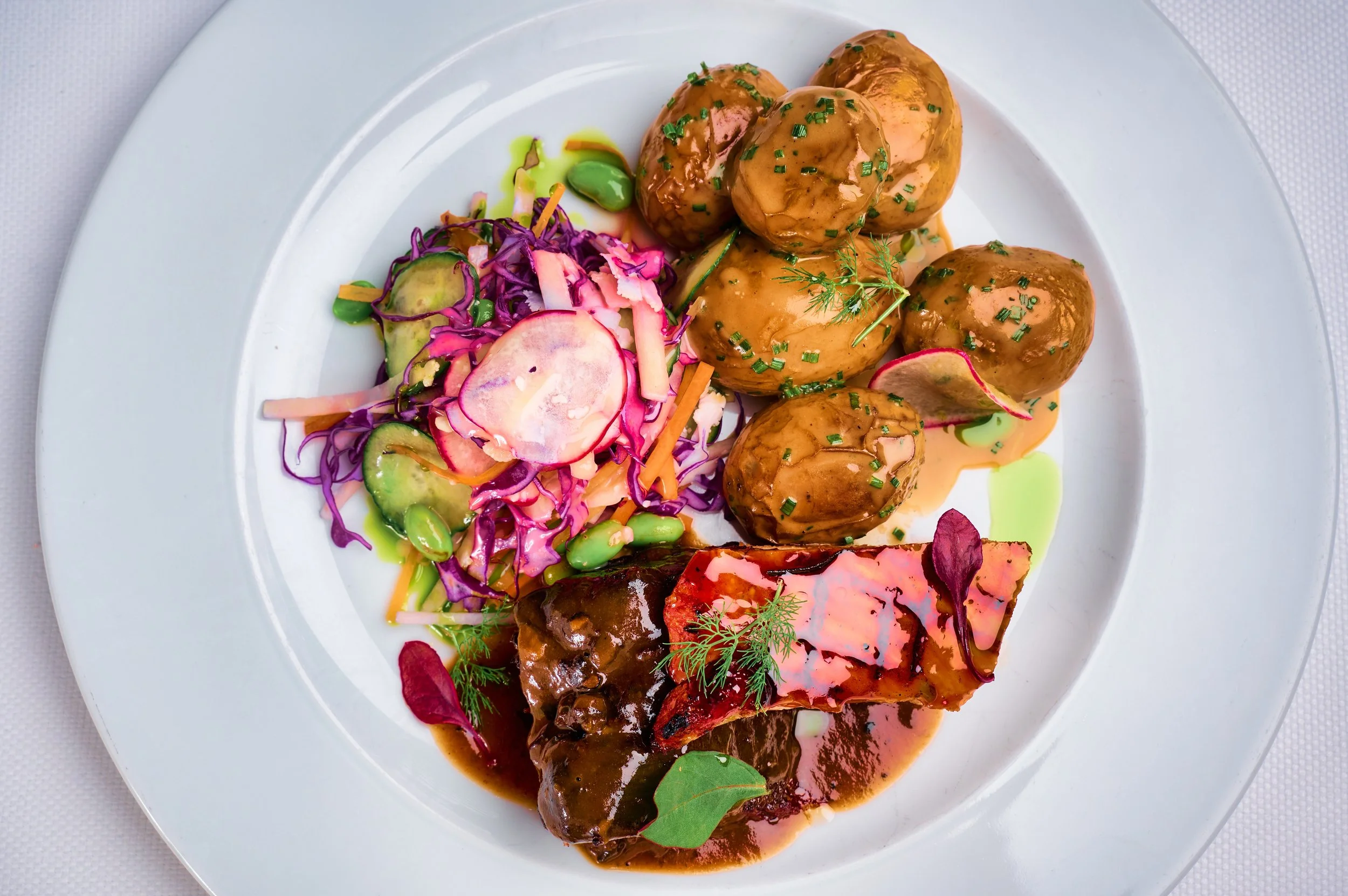 A white plate with meatballs covered in brown gravy, garnished with chopped chives, alongside a mixed salad with purple cabbage, sliced radishes, and cucumbers, with drizzles of green sauce.