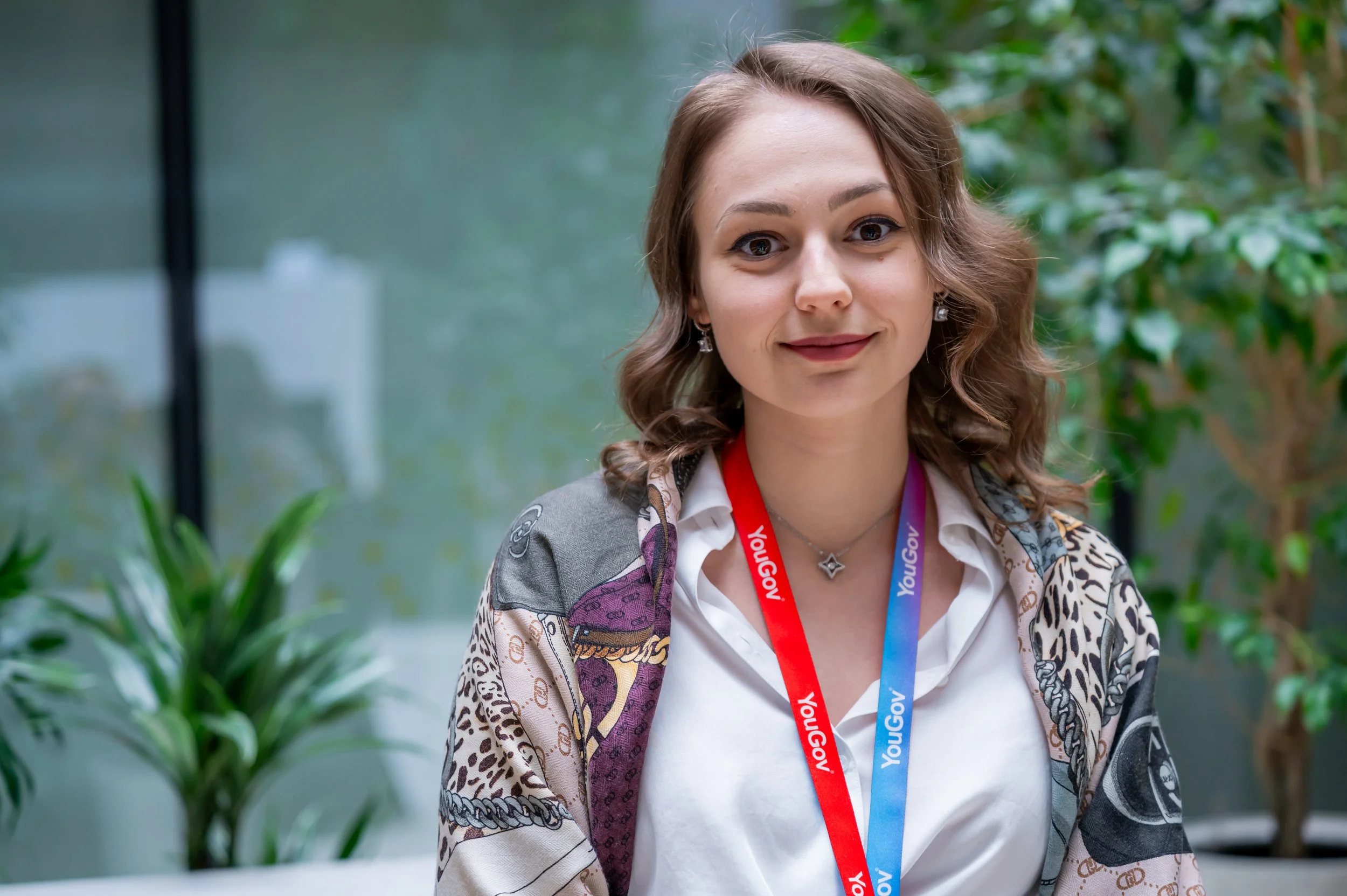 A woman with light brown wavy hair wearing a white blouse and a beige patterned jacket, standing indoors with green plants in the background. She has a red and blue lanyard around her neck that says 'YoungGov'.