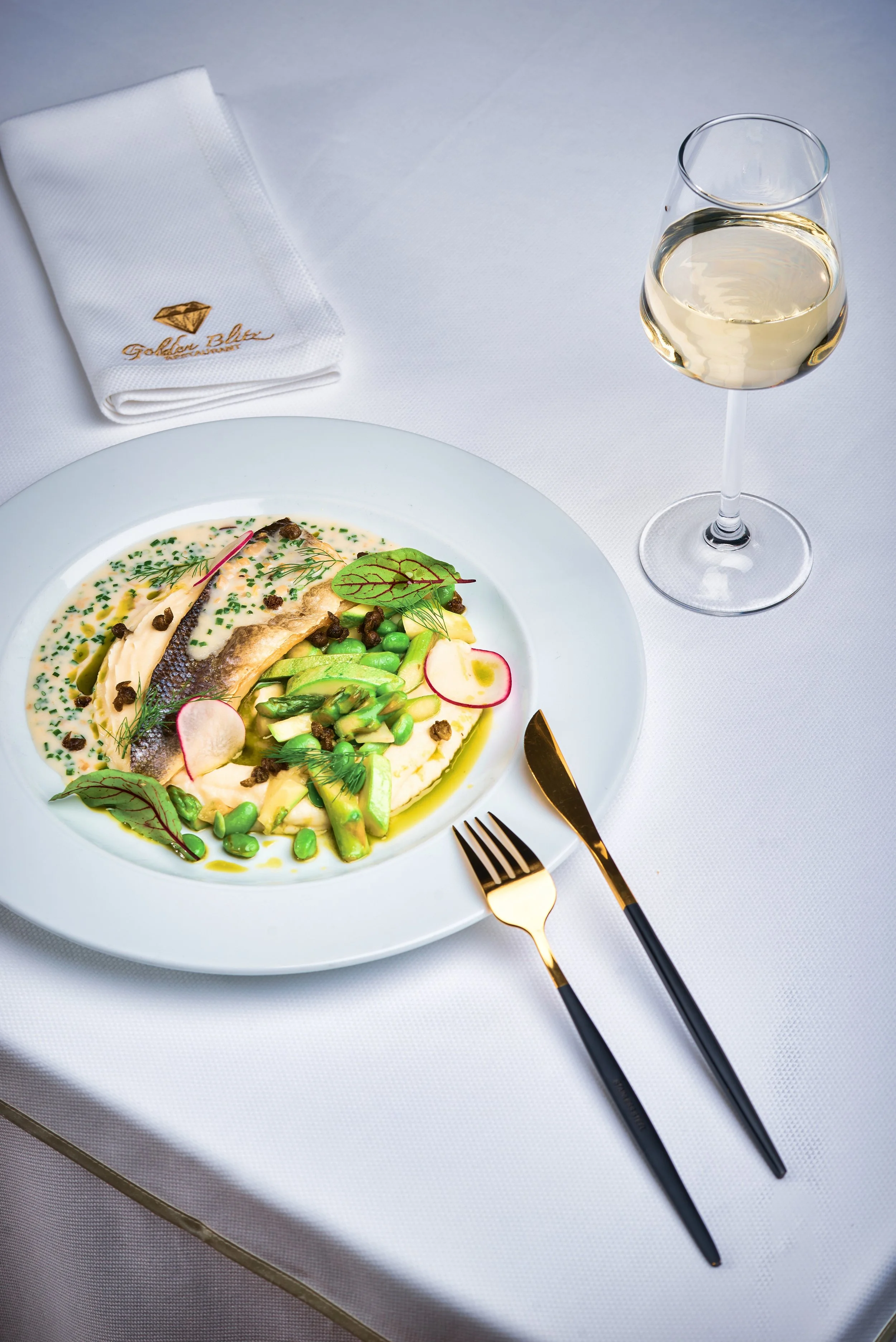 A plate of fish with vegetables, garnished with herbs and sauce, placed on a white table with black and gold utensils. There is a glass of white wine and a paper napkin with gold embroidery reading "Golden Blue".