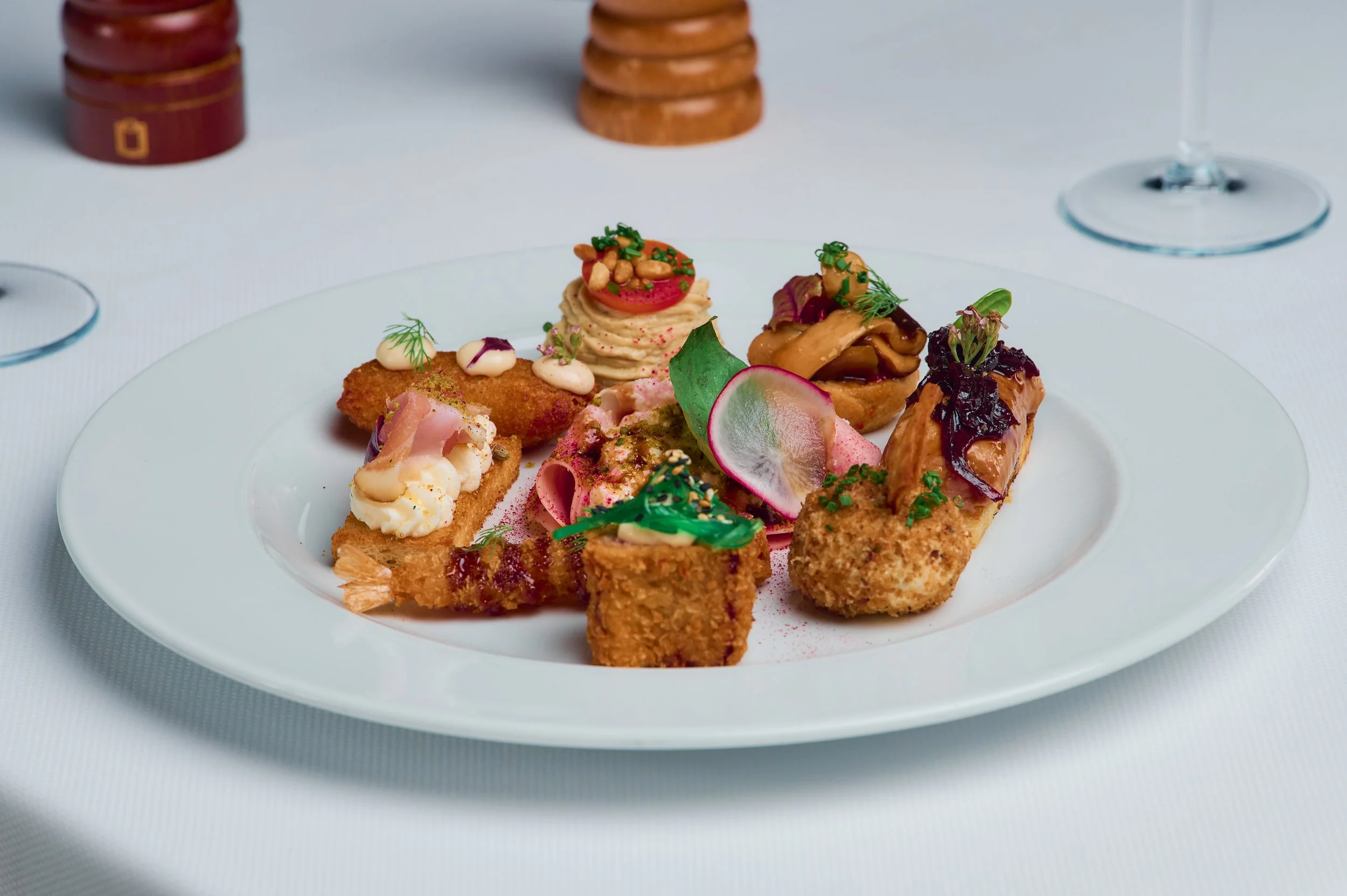 A white plate with various gourmet appetizers, including croquettes, a slice of radish, garnished with herbs and sauces, with glasses and salt and pepper shakers in the background.