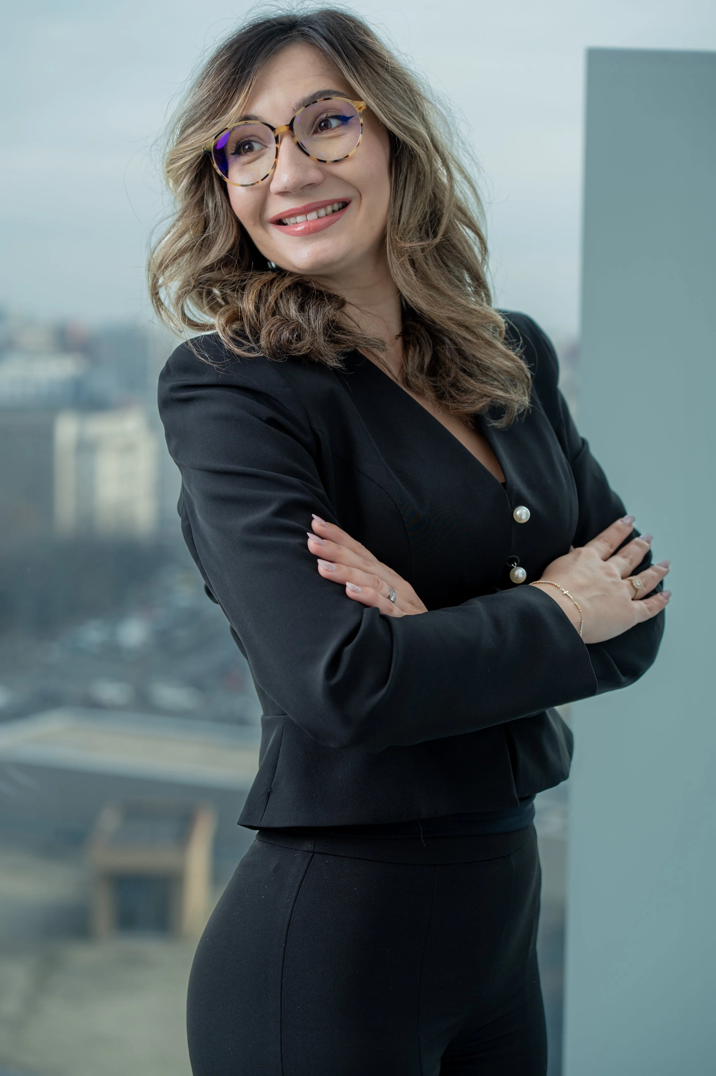 A woman in professional attire with cross-arms, wearing glasses, smiling, in an office setting with city buildings in the background.