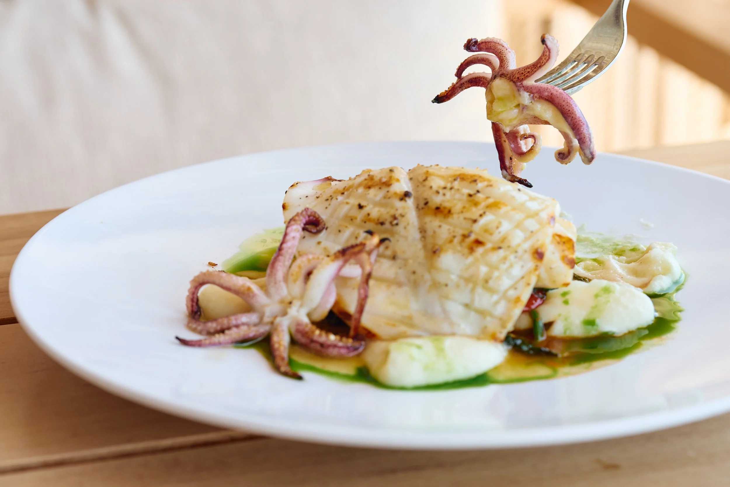 A plate with cooked fish, topped with small octopus tentacles, garnished with green sauce, served on a white plate.