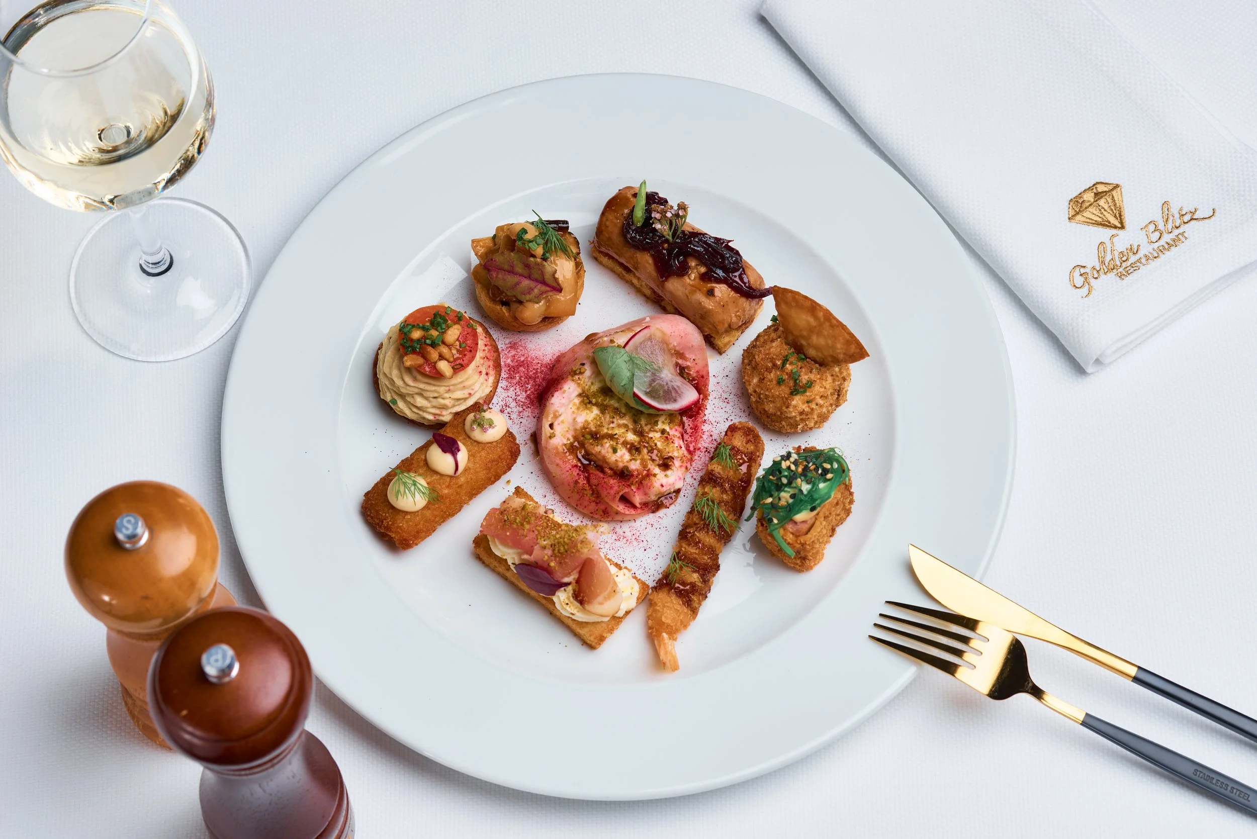 A white round plate with assorted gourmet appetizers, including small sandwiches, canapés, and spreads, is placed on a white tablecloth. To the left, there are three bottles of sauces or condiments with metal caps. To the right, there is a white napk