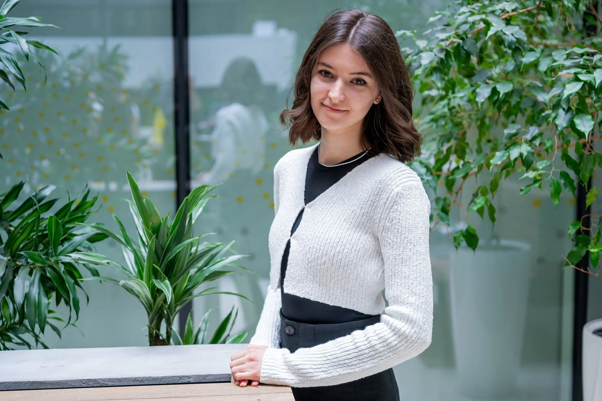 Young woman with shoulder-length brown hair, wearing a white cardigan over a black top, standing near green plants indoors.