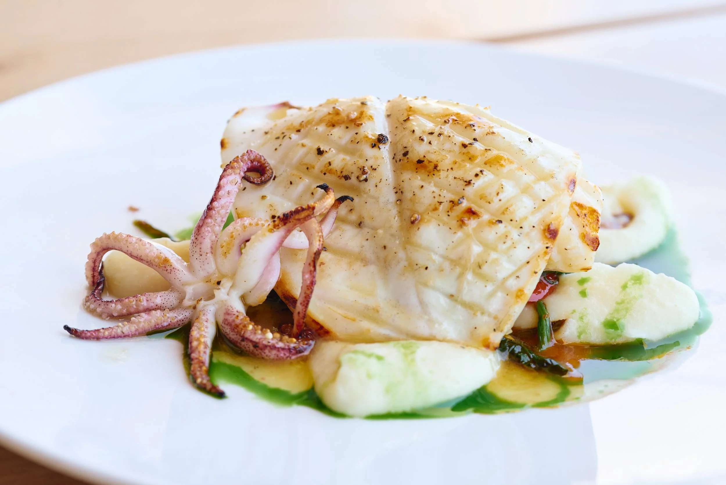 A close-up of a gourmet seafood dish featuring a cooked fillet of fish topped with herbs and black pepper, accompanied by small squid and colorful vegetable garnishes on a white plate.