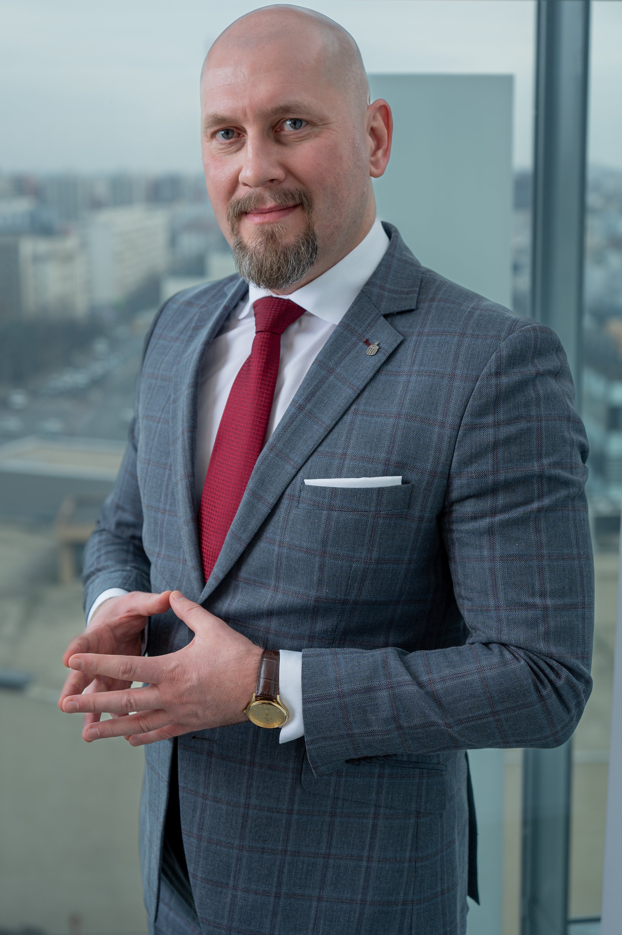 A man in a gray checkered suit with a red tie, standing in an office with large windows showing city buildings. He has a beard and is wearing a wristwatch and a lapel pin. Someone's hands are adjusting his suit jacket.