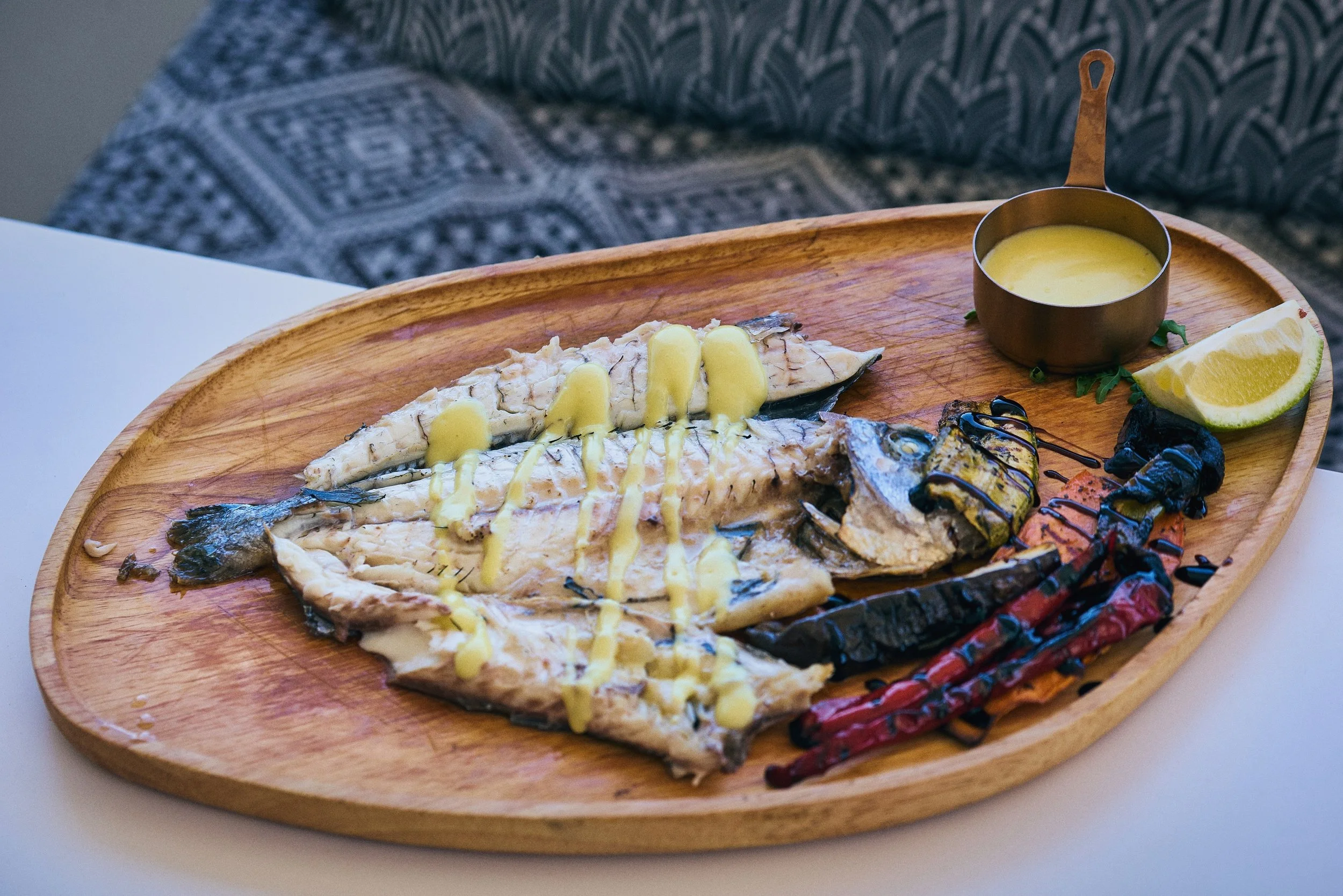 Grilled fish fillet topped with lemon sauce on a wooden platter with lemon wedges and charred vegetables, served with a side of sauce in a small copper bowl.