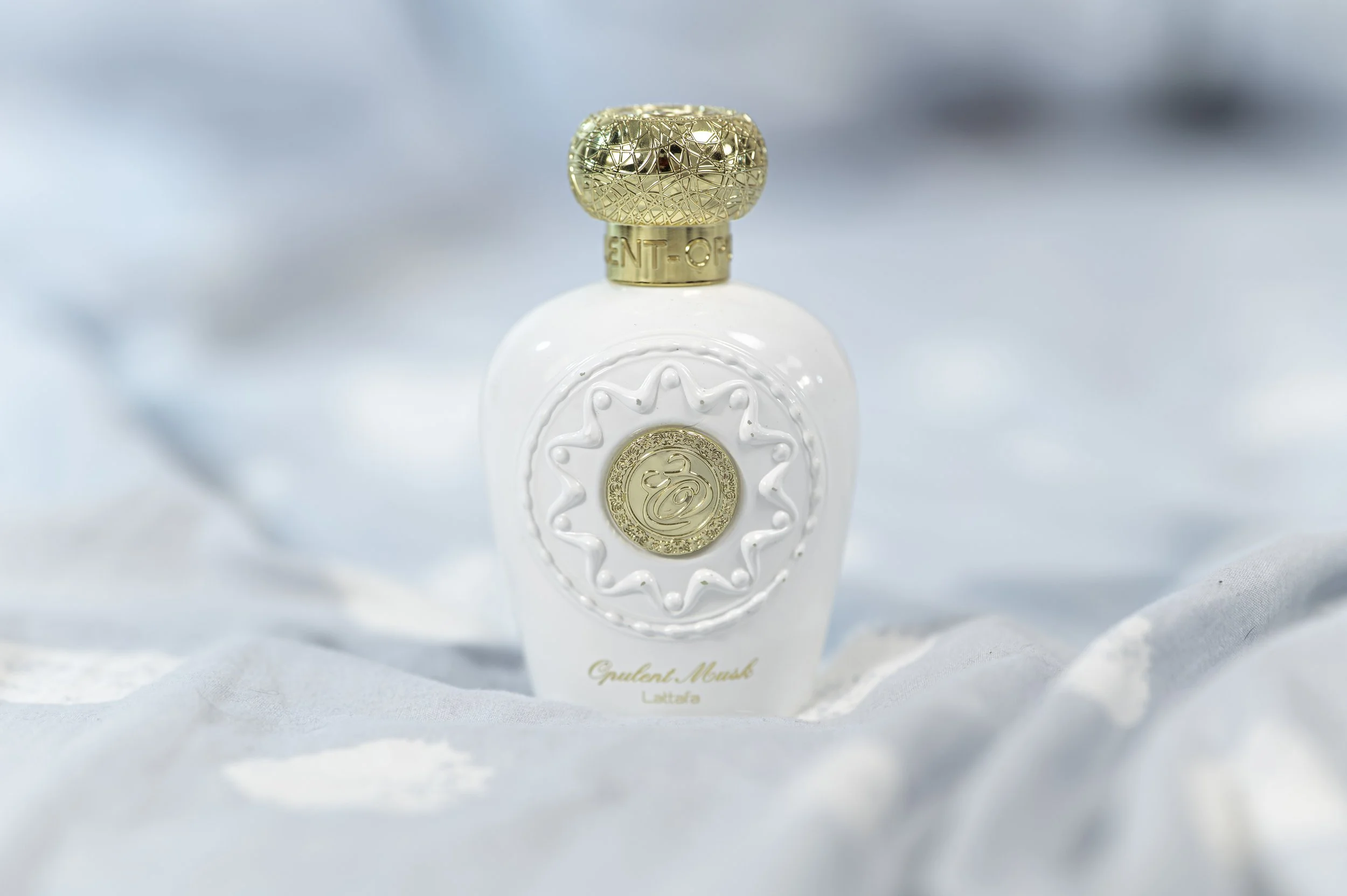 A white perfume bottle with a gold cap, decorated with embossed gold accents and text, against a soft, light blue background.