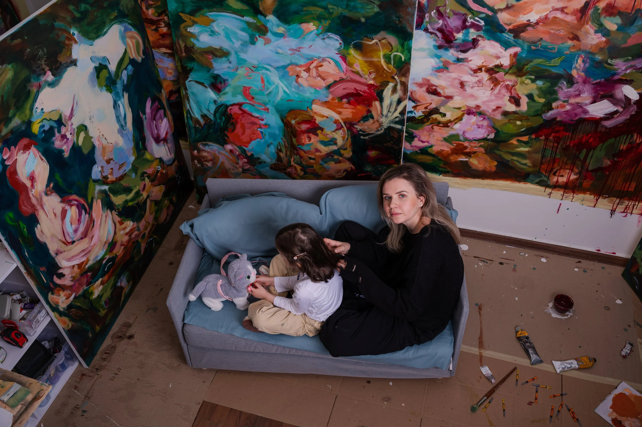 A woman and a young girl sitting on a bed surrounded by colorful, abstract floral paintings in an art studio. The girl is playing with a stuffed animal.