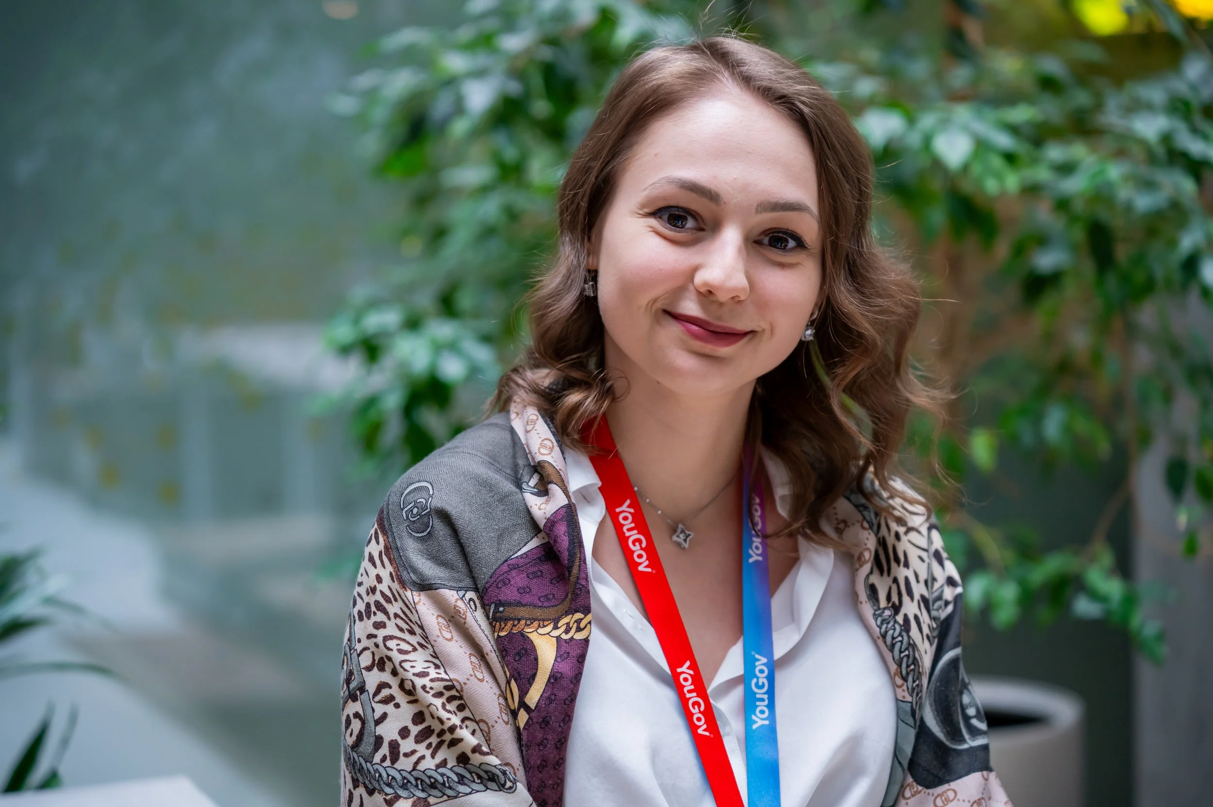A woman with brown hair in loose waves, smiling, wearing a patterned jacket with animal prints, white shirt, and a red and blue lanyard, standing in front of green foliage.