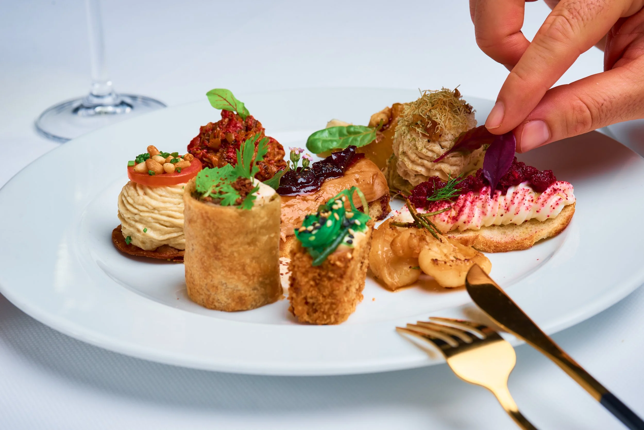 A white plate with an assortment of appetizers including stuffed mushrooms, a small cone-shaped pastry, a creamy dip with a tomato slice, a piece of flatbread with pink seasoning, and other small garnished bites. A person's hand is adding a garnish t
