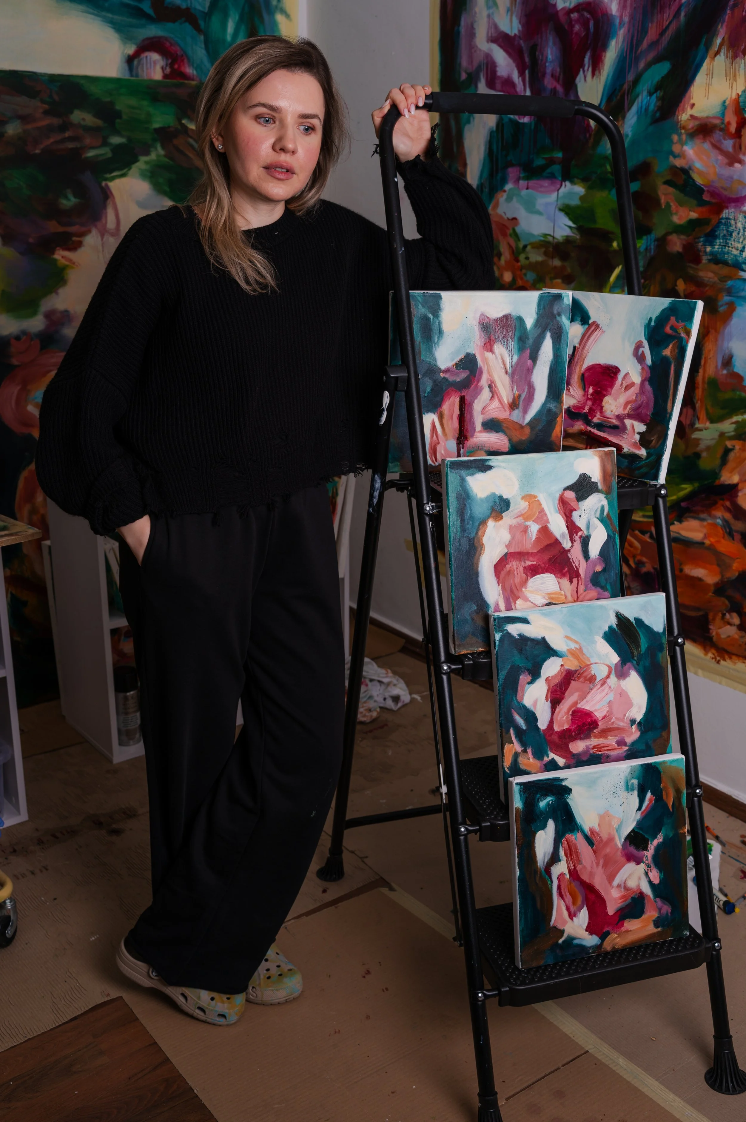 A woman standing in an art studio holding a black step ladder with multiple abstract paintings on it, and a large colorful abstract painting in the background.
