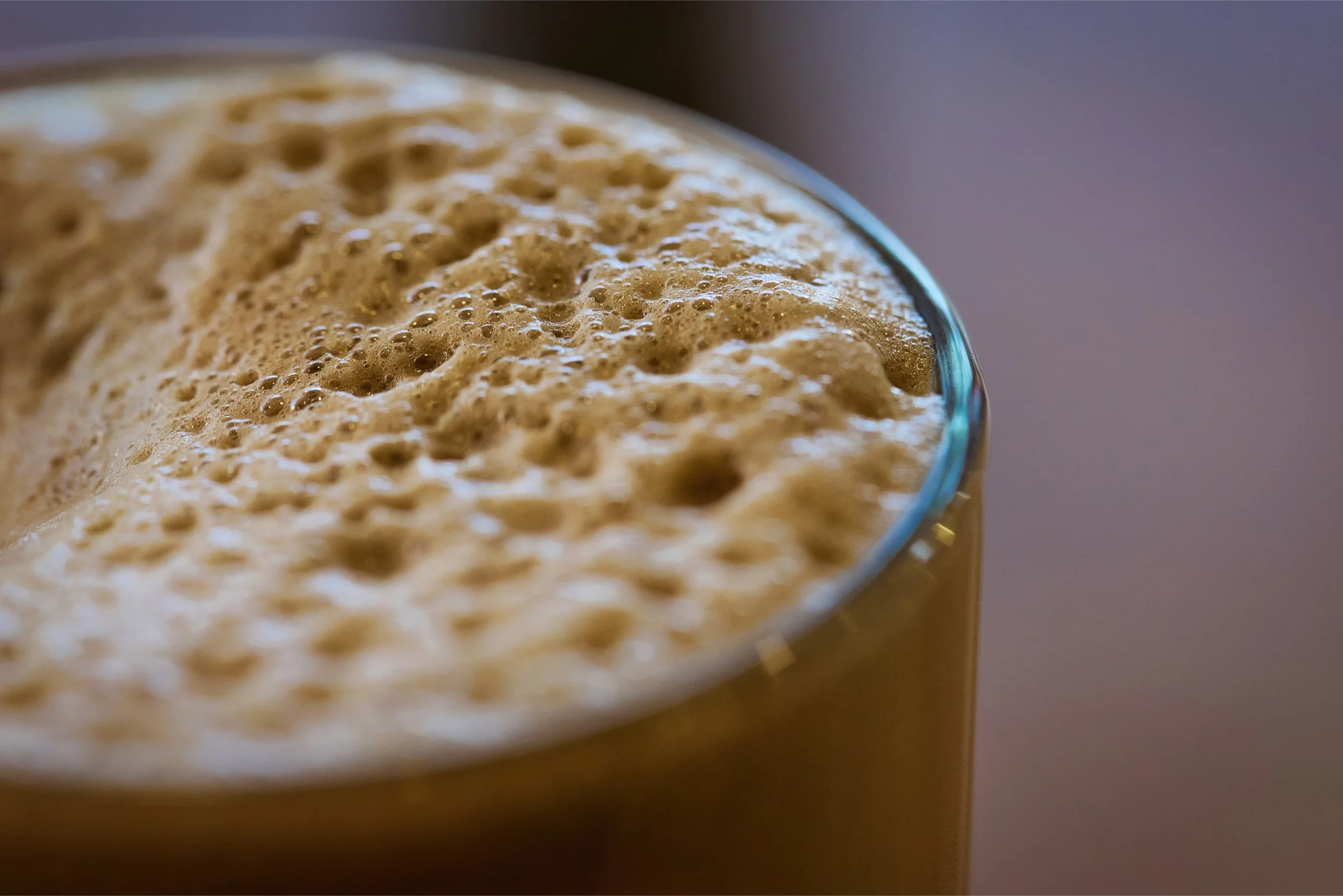 Close-up of a glass filled with frothy coffee or tea with foam bubbles on top, against a plain background.