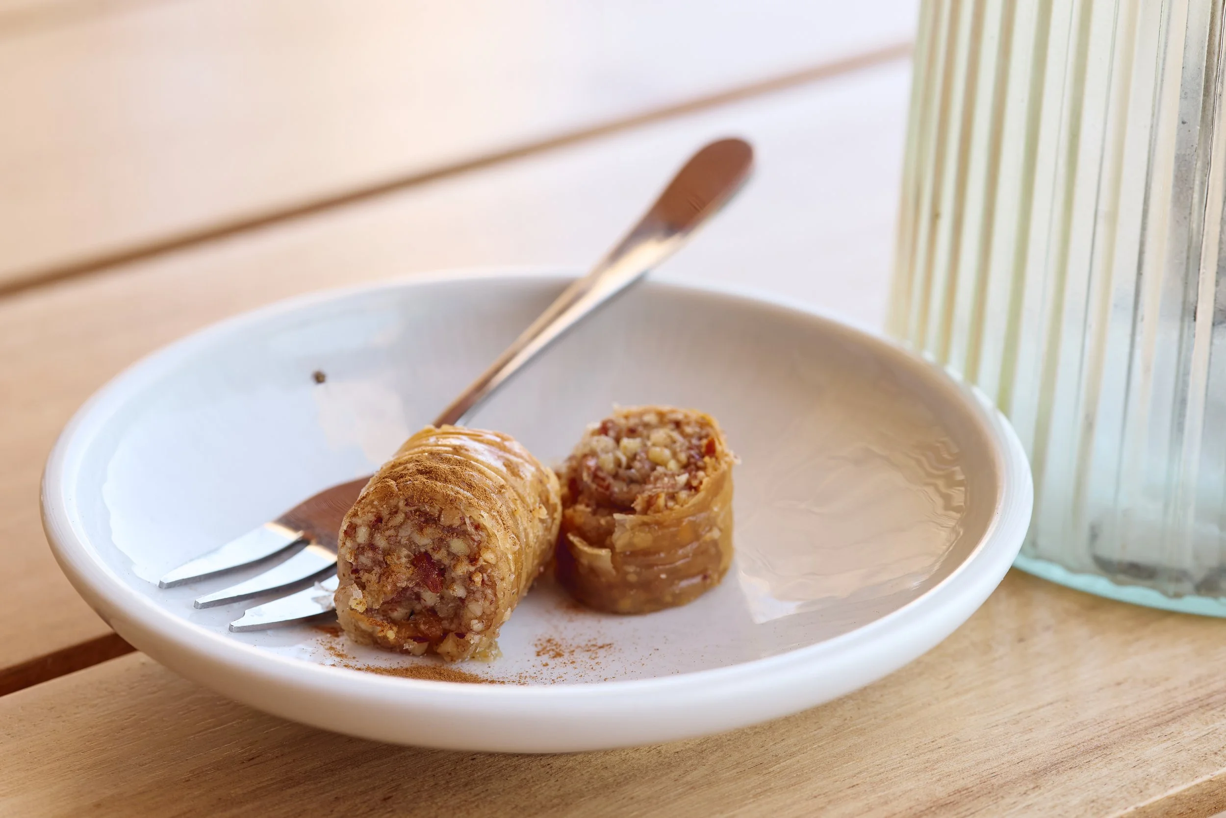 Close-up of apple cinnamon roll split in half on a white plate with a fork on side, next to a glass of water, on a wooden surface near a window.