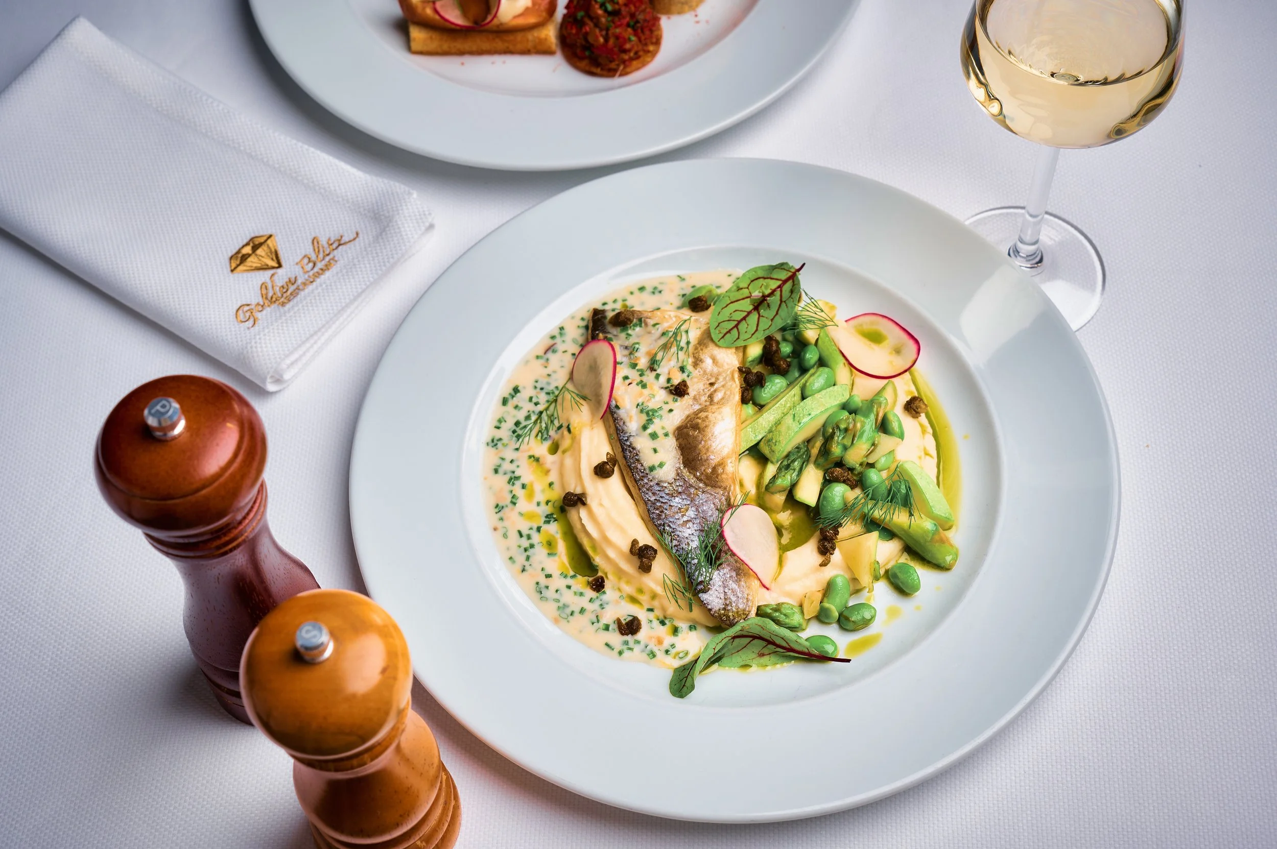 A white plate containing a fish fillet with a creamy sauce, garnished with radish slices, microgreens, capers, and green peas. A glass of white wine and a white napkin with gold embroidery are also visible on a white tablecloth.