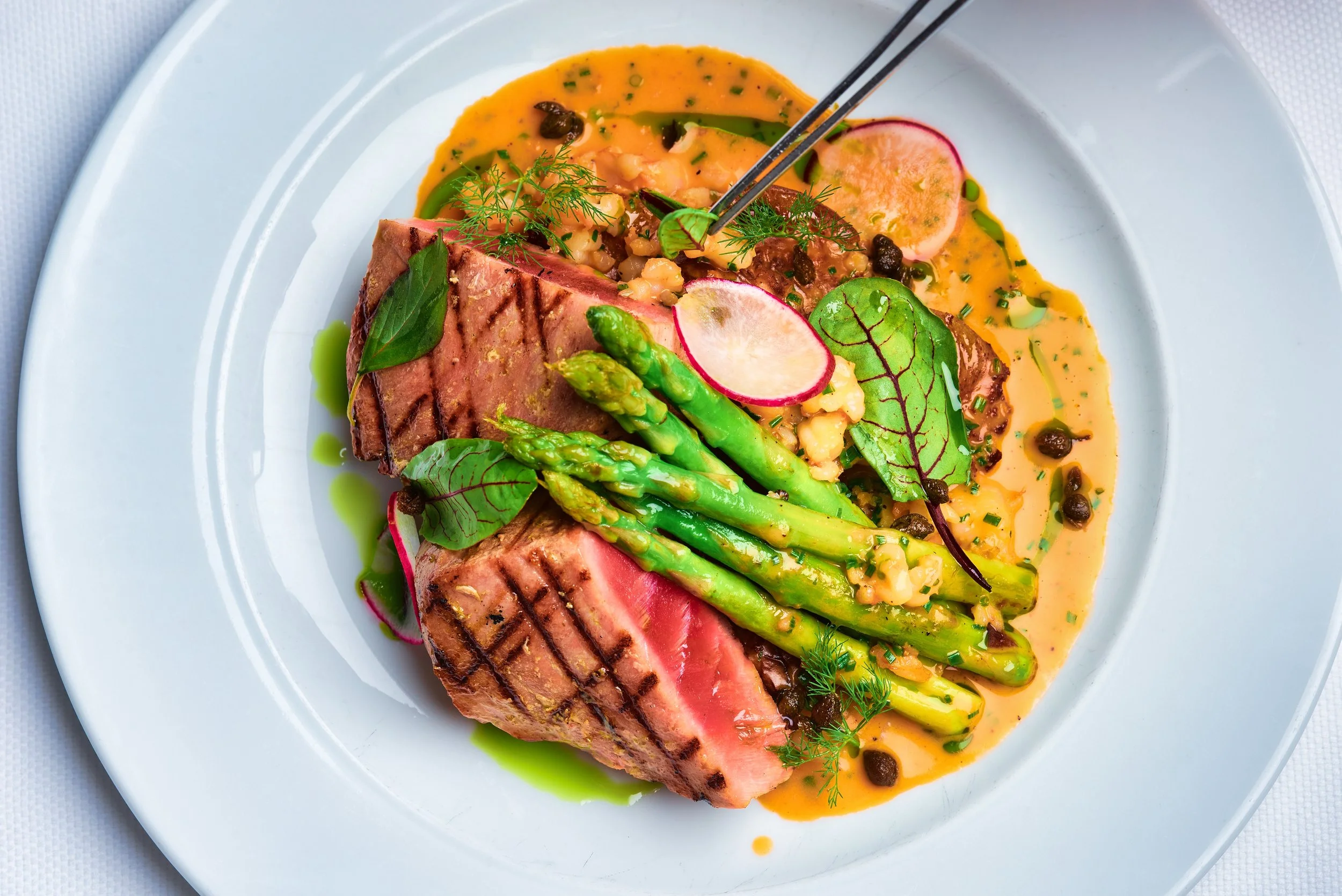 A white plate with a medium-rare grilled steak, grilled asparagus, radish slices, microgreens, and a colorful sauce with herbs and peppercorns.