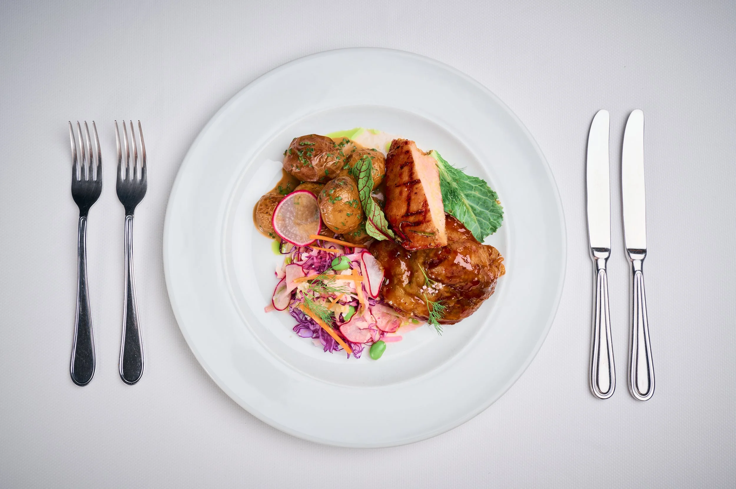 White plate with grilled salmon, small potatoes, and colorful salad, on a white tablecloth with silverware on each side.