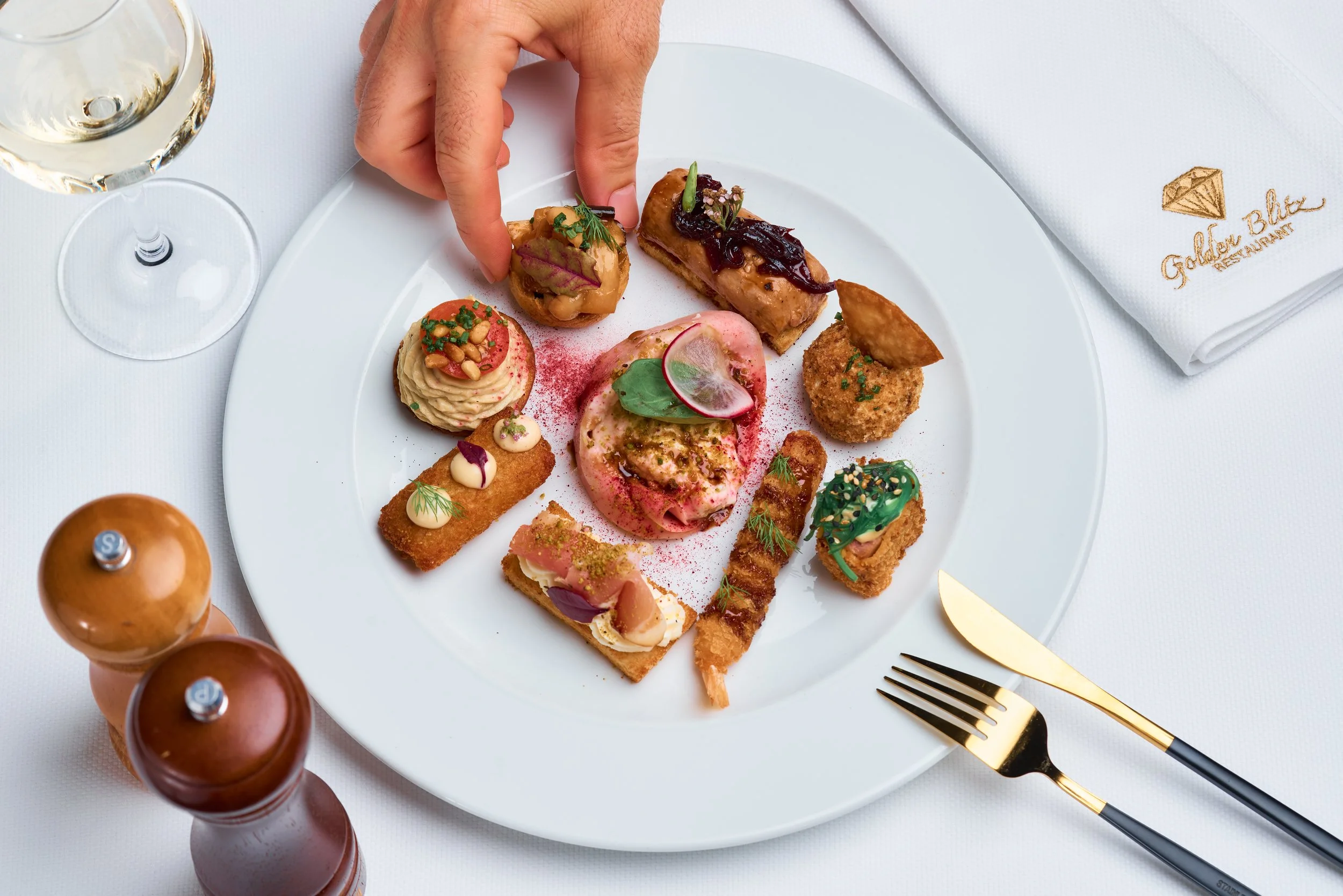 A white plate with a variety of small gourmet appetizers and a glass of white wine on a white tablecloth at a restaurant. Gold and black cutlery, napkin with embroidery.