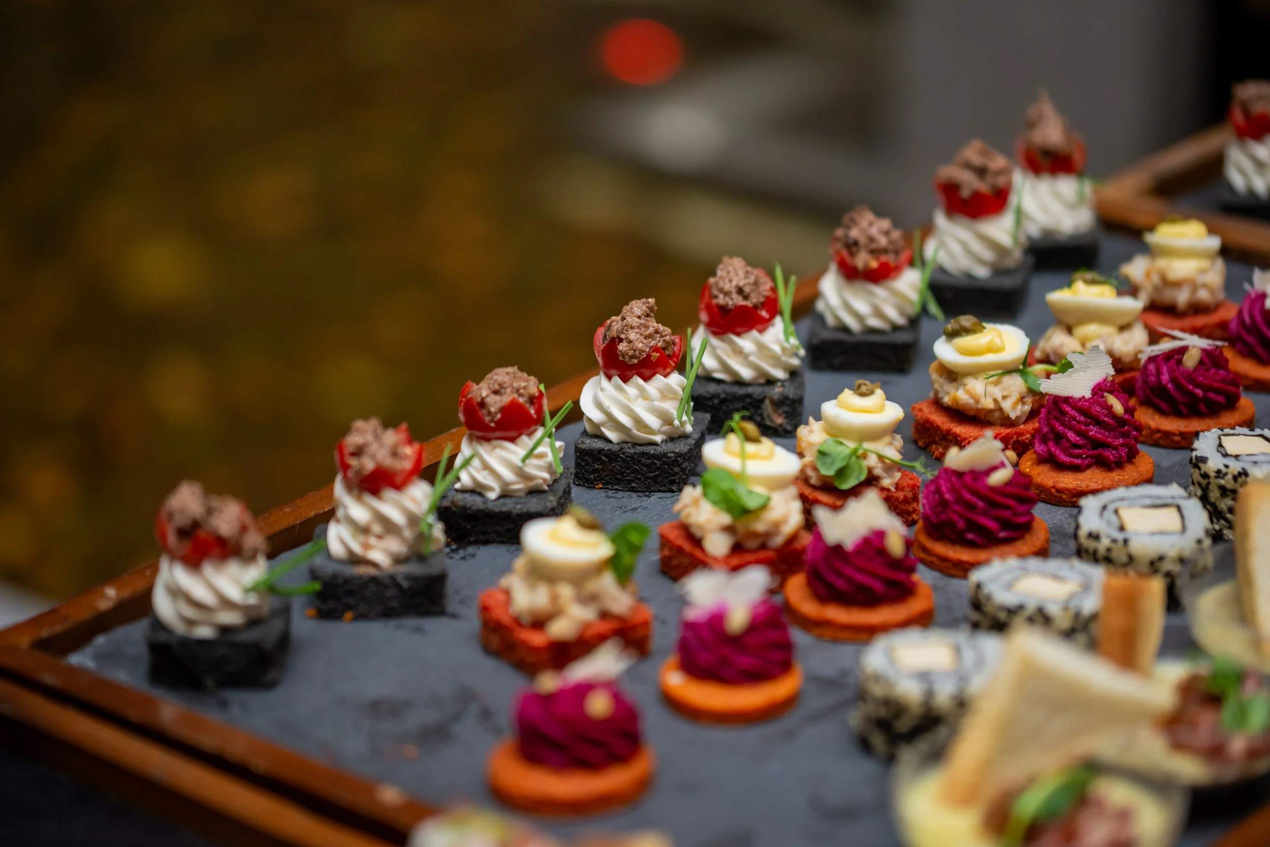 Assorted small appetizers on a black slate platter, including sushi with various toppings, garnished with herbs and vegetables.