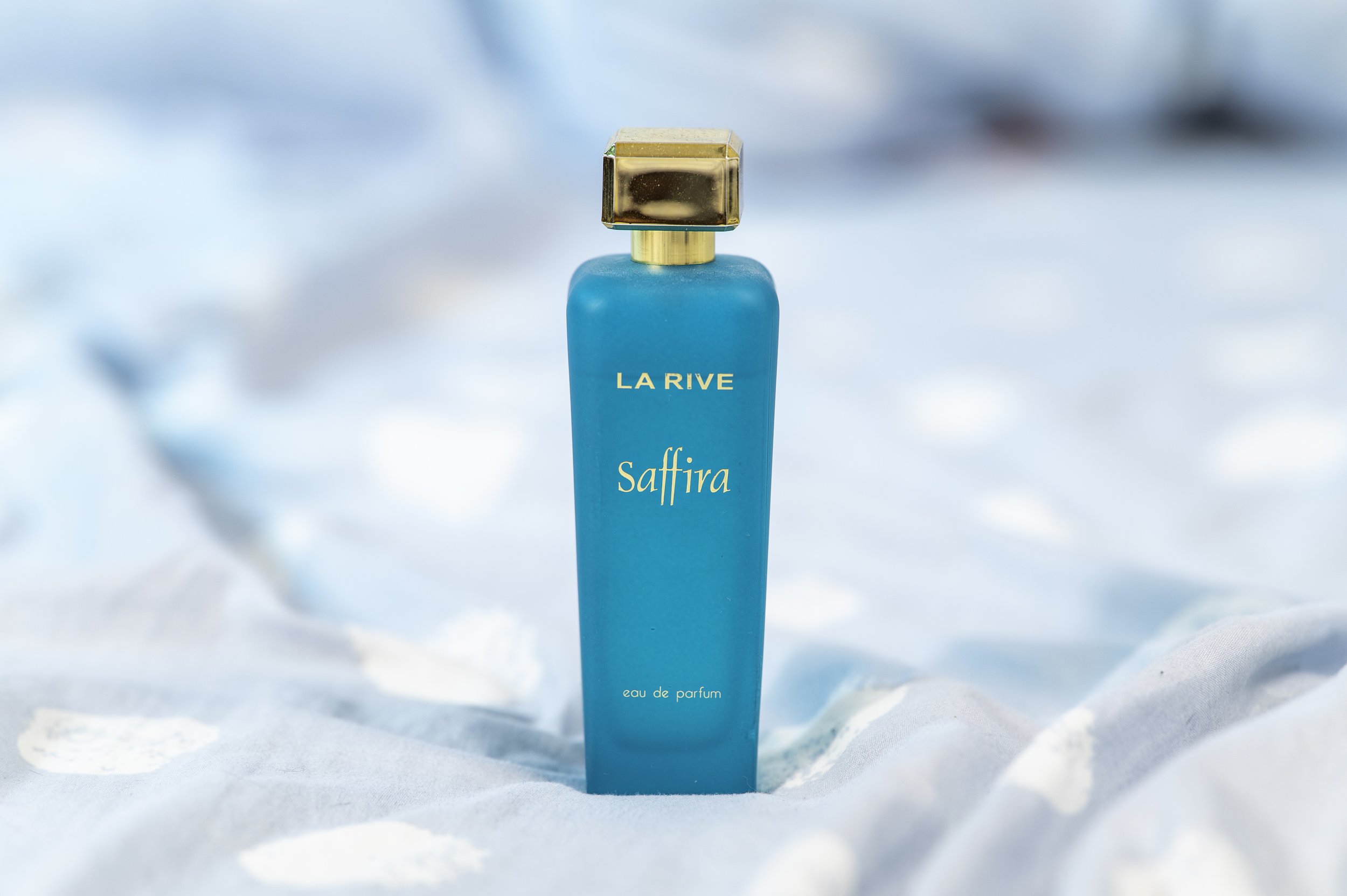 A turquoise perfume bottle with gold accents and a square gold cap, labeled 'LA RIVE Saffira', against a soft, blurry background.