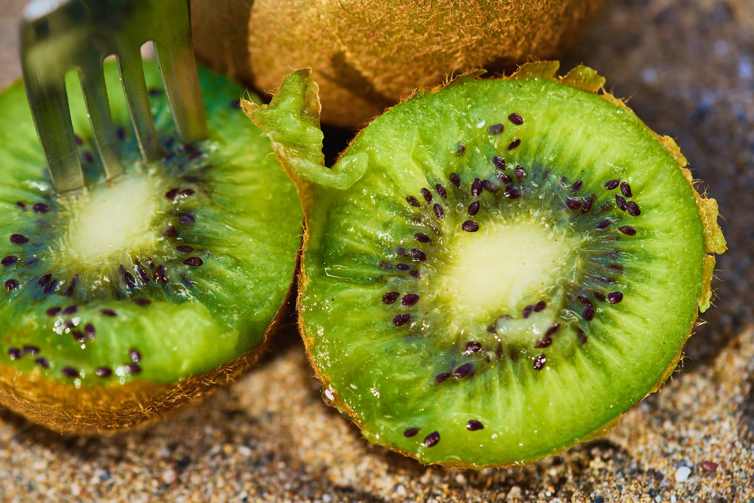 Close-up view of a halved kiwi fruit on sandy ground, showing green flesh with black seeds.