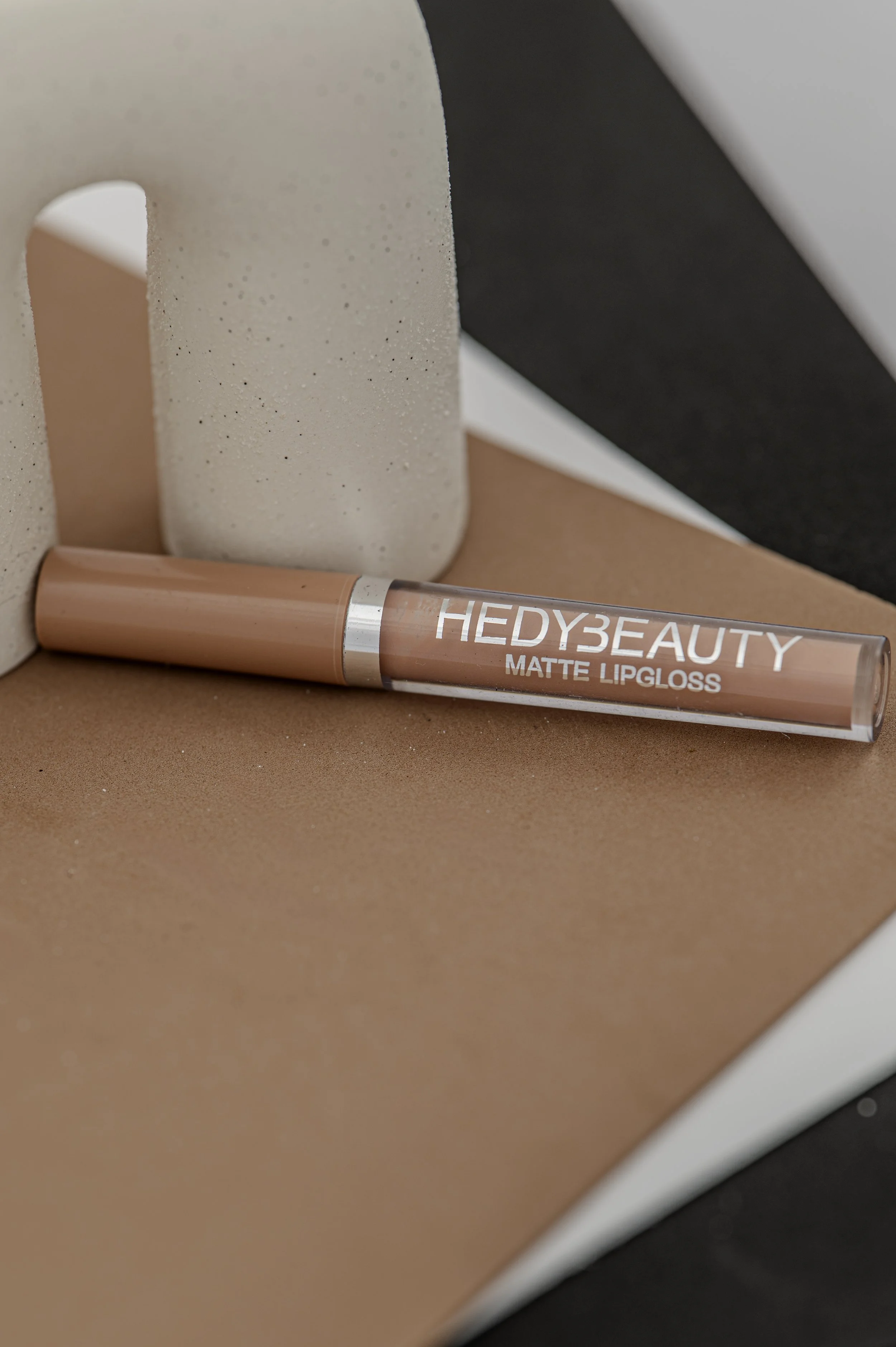 Close-up of Hedy Beauty matte lip gloss tube lying on a peach-colored surface with a geometric background of beige, black, and white shapes.