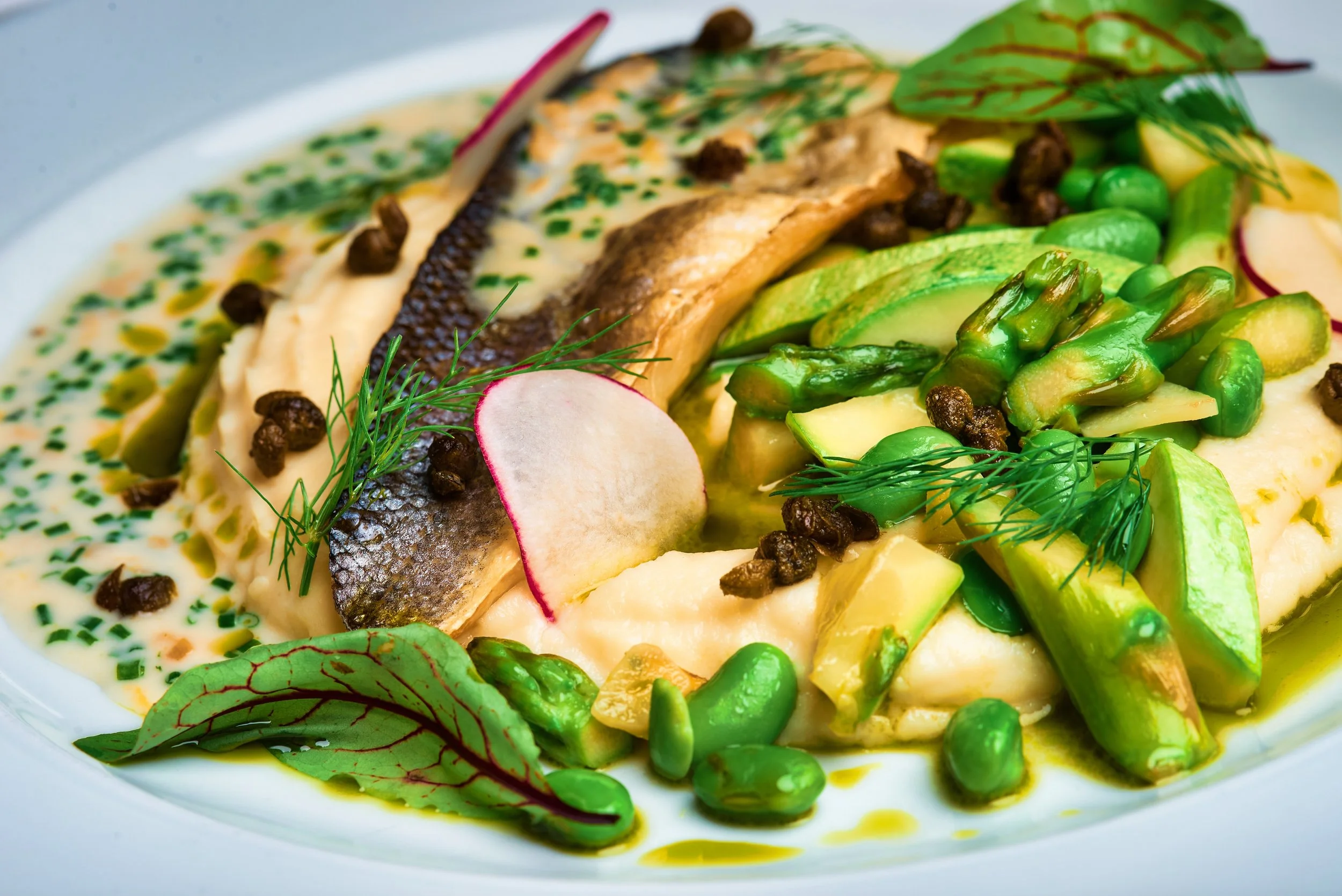 A dish featuring a piece of cooked fish with crispy skin, served on a bed of creamy mashed potatoes, garnished with sliced radish, fresh dill, and parsley, with asparagus spears and green peas alongside.