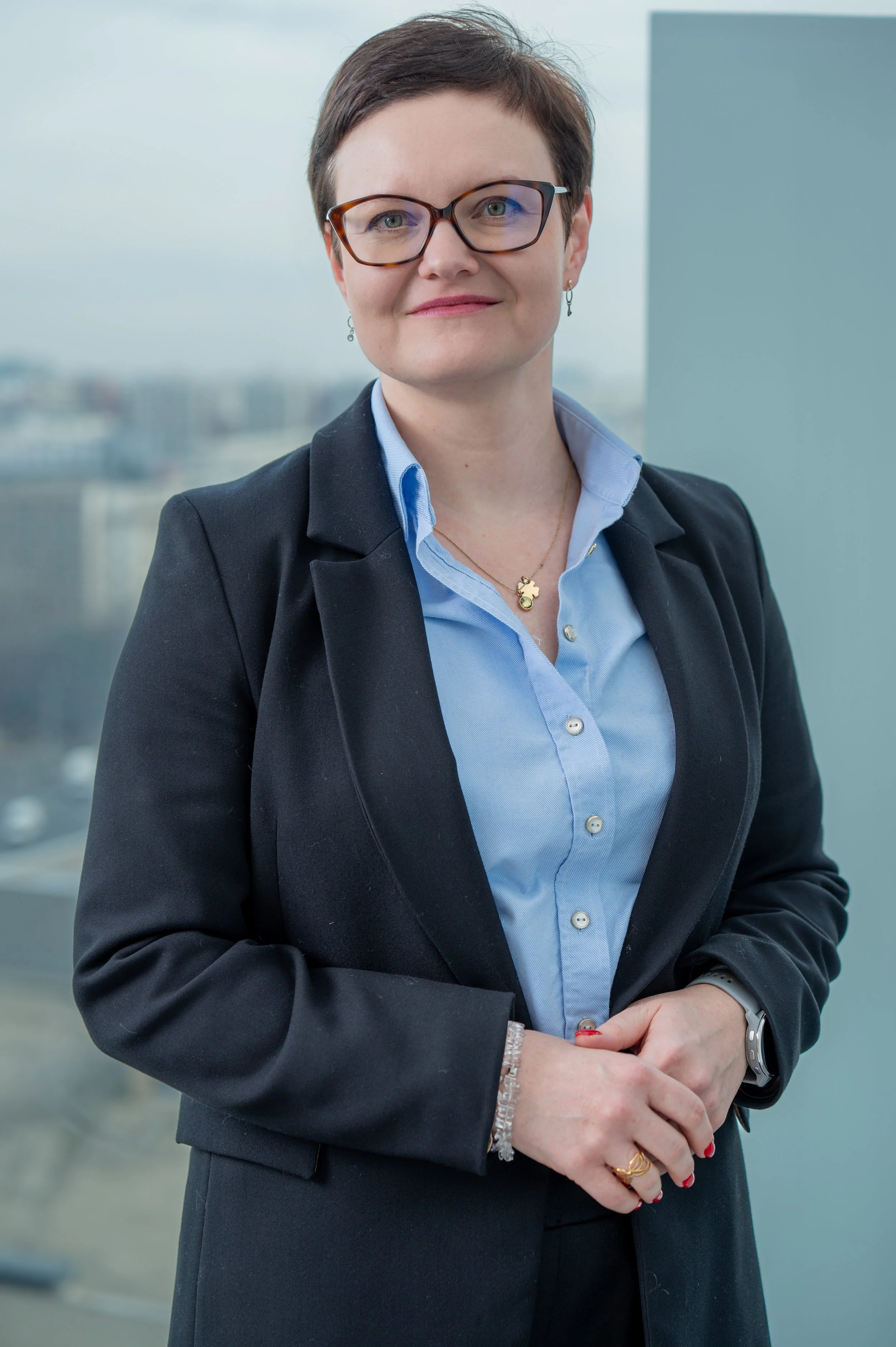 A professional woman in a business suit and glasses standing with her hands clasped in front of a window overlooking a cityscape.