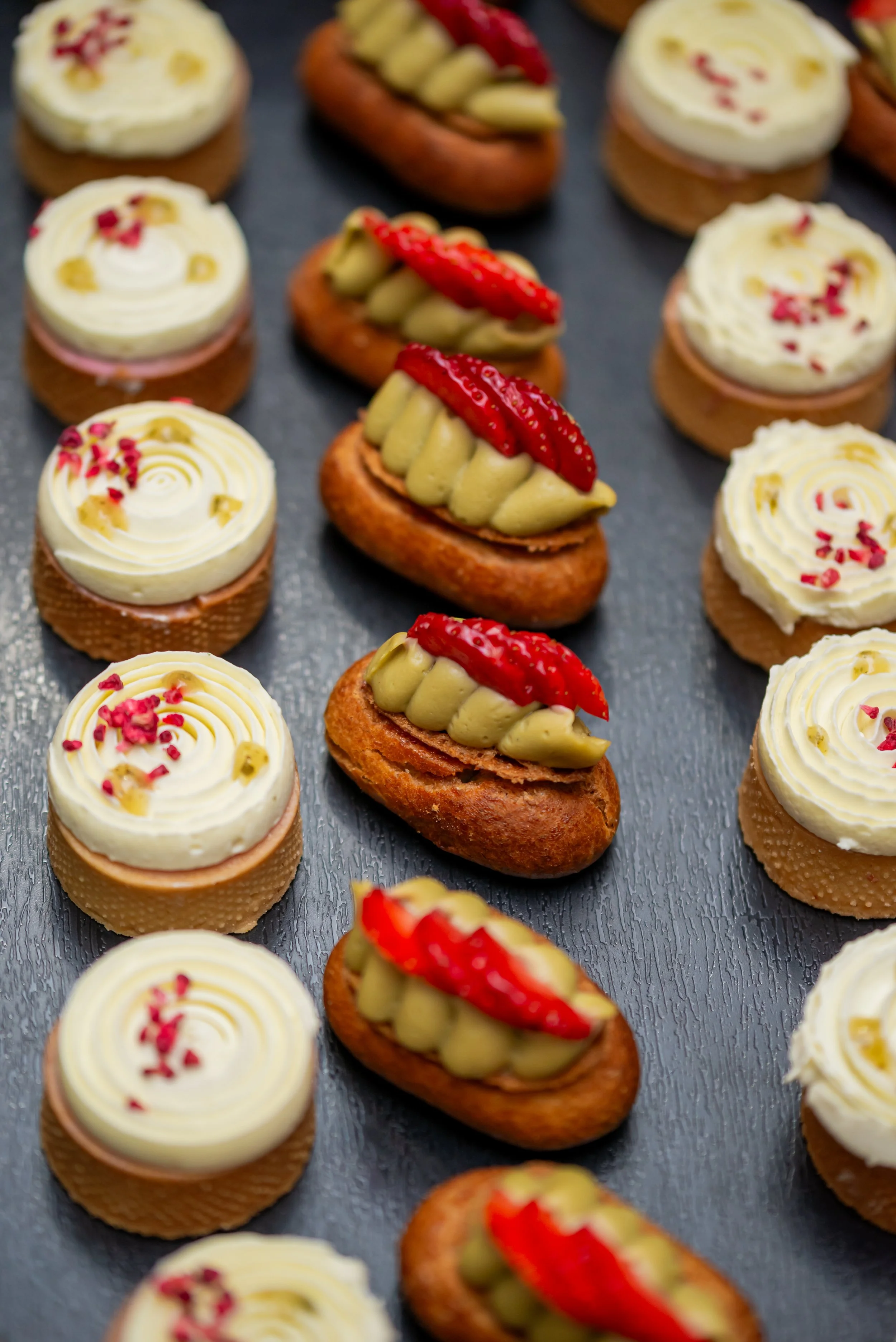 Close-up view of assorted pastries, including small cupcakes with swirled white frosting decorated with pink and yellow sprinkles, and elongated éclairs topped with sliced strawberries and green filling, arranged on a dark surface.
