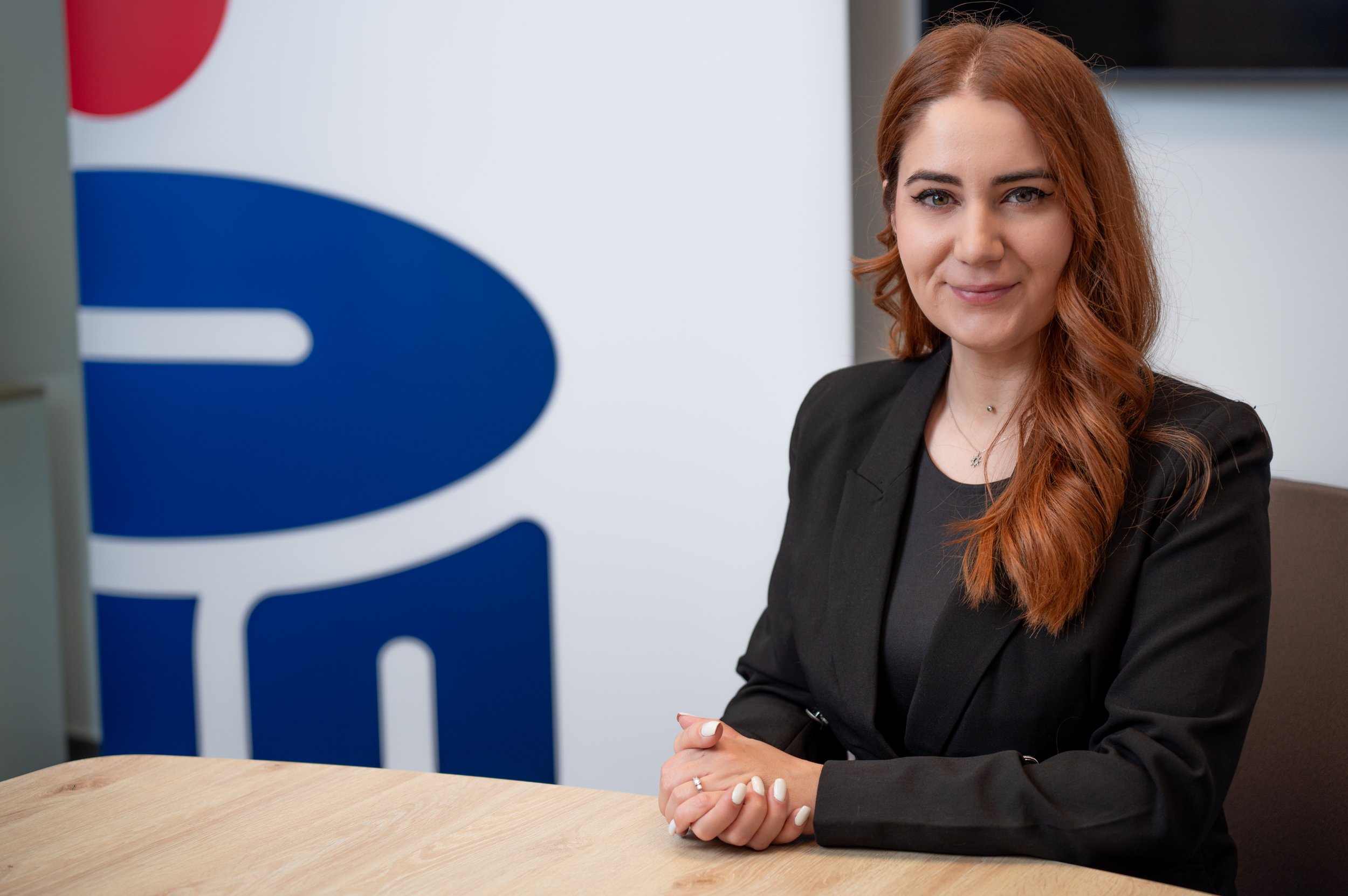 A woman with long red hair and a black blazer sitting at a wooden table in a professional setting, with a blurred corporate logo or sign in the background.