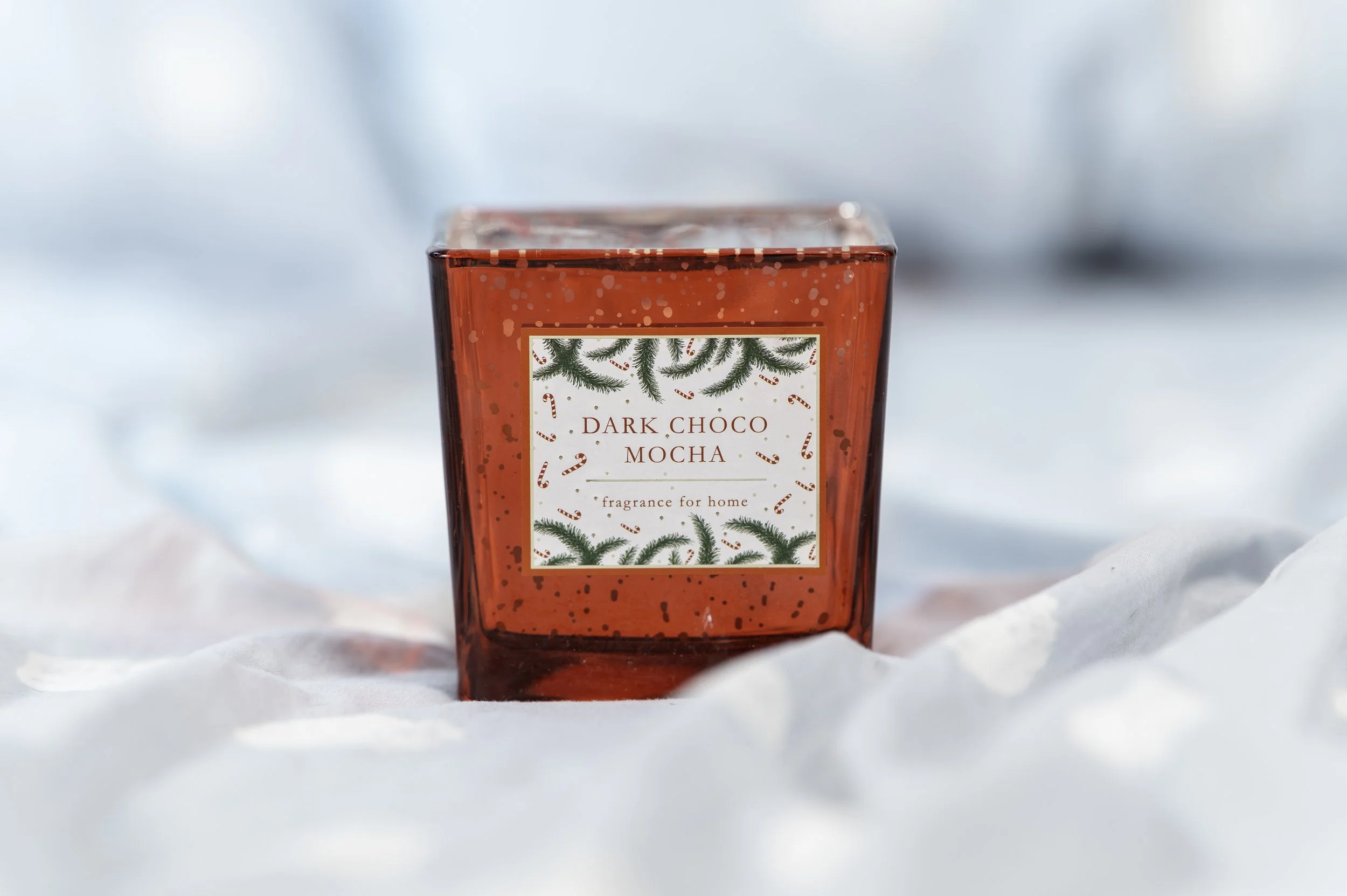 A red glass candle holder with a festive label reading 'Dark Choco Mocha, fragrance for home,' surrounded by holiday decorations and set on a snowy surface.