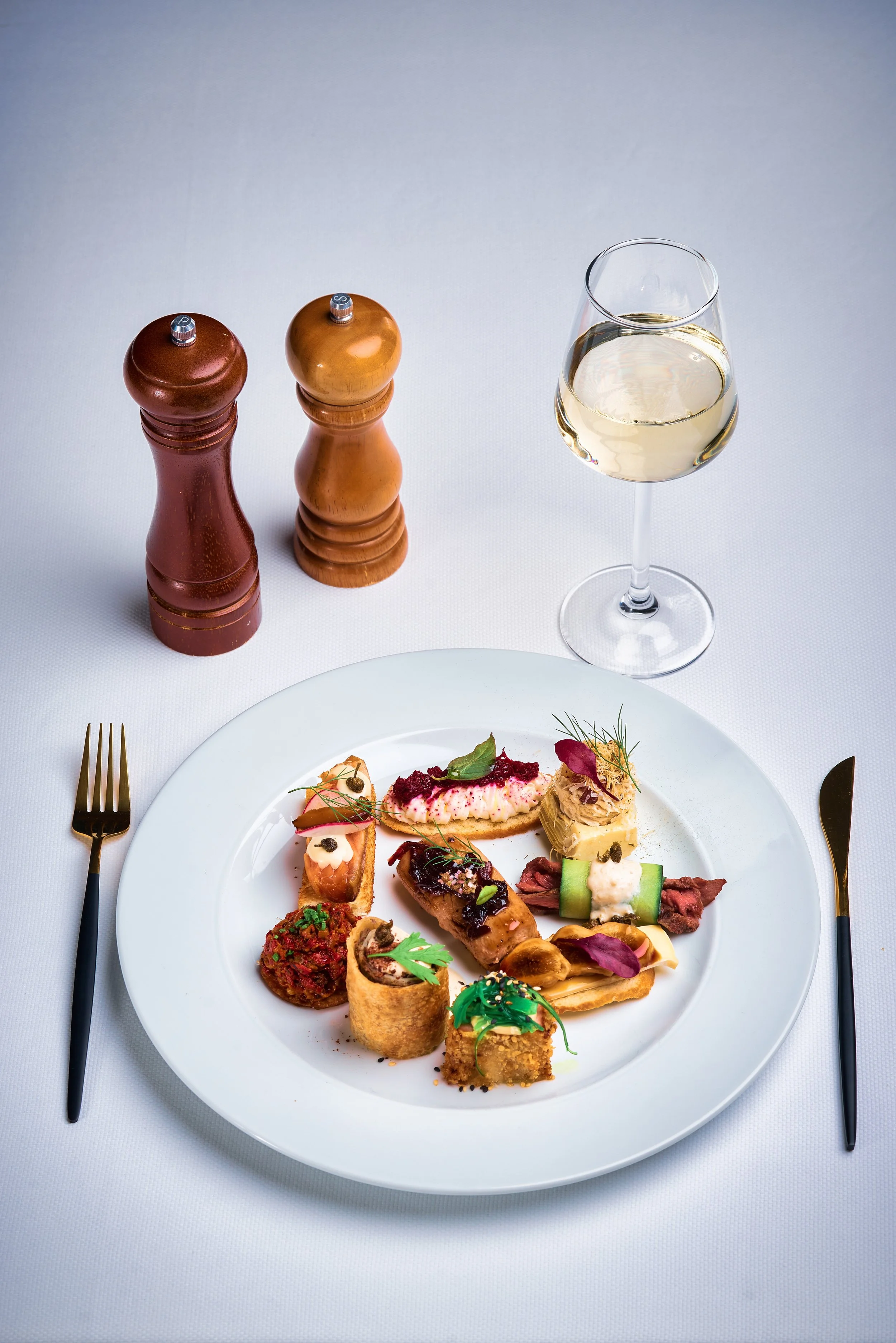 A white plate with assorted appetizers on a table set with a fork and knife, a glass of white wine, and two salt and pepper shakers.