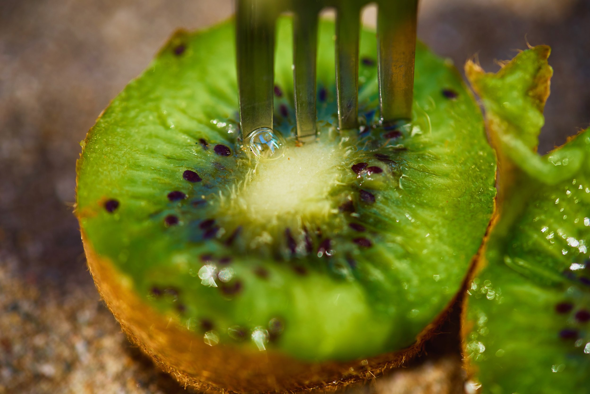 A close-up of a sliced kiwi with a fork inserted into the flesh of the fruit, showing its green interior and black seeds.