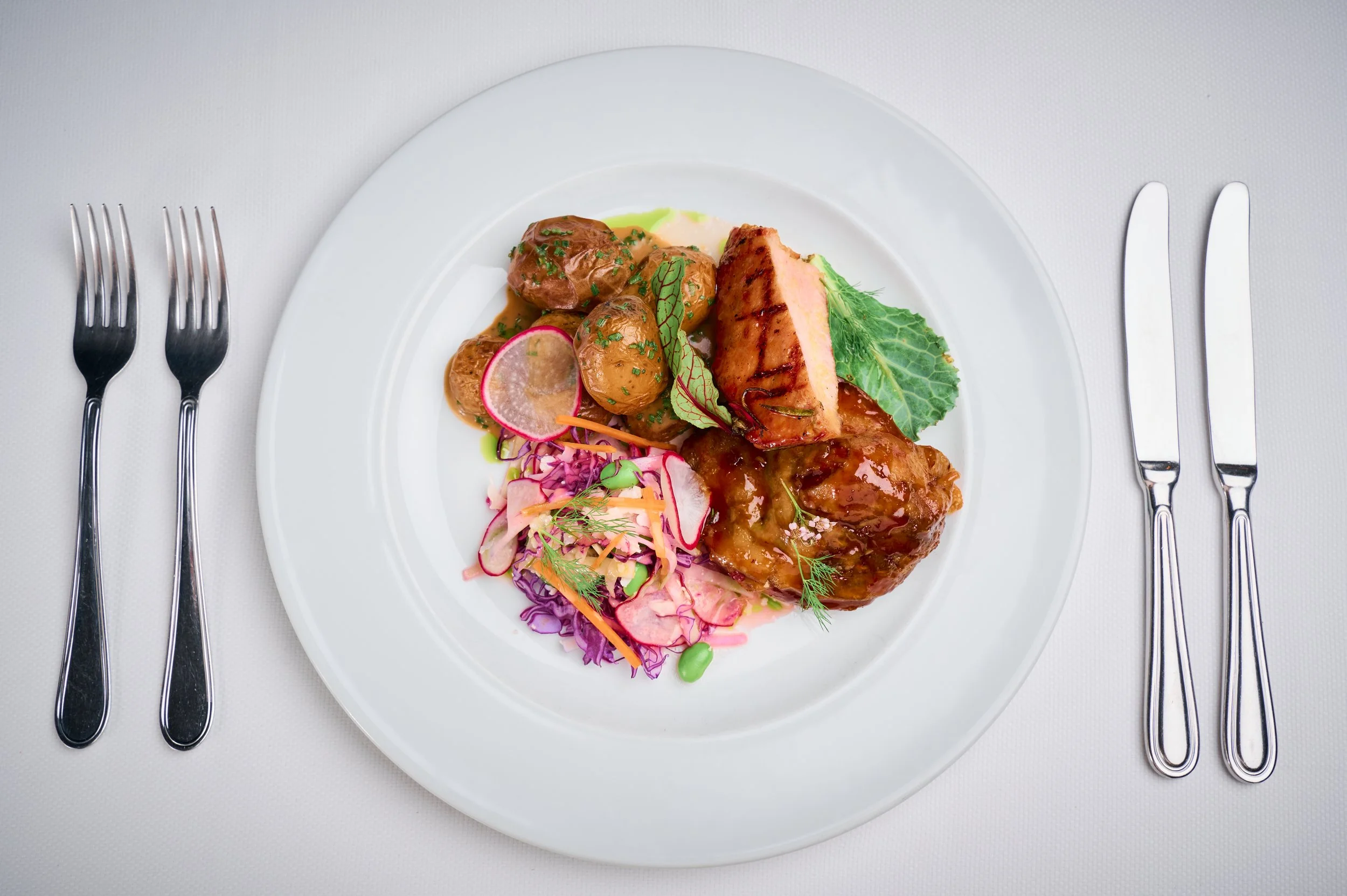 A white plate with grilled salmon, roasted potatoes with herbs, and colorful salad with radish, carrots, and lettuce, garnished with herbs, on a white tablecloth with silverware on each side.