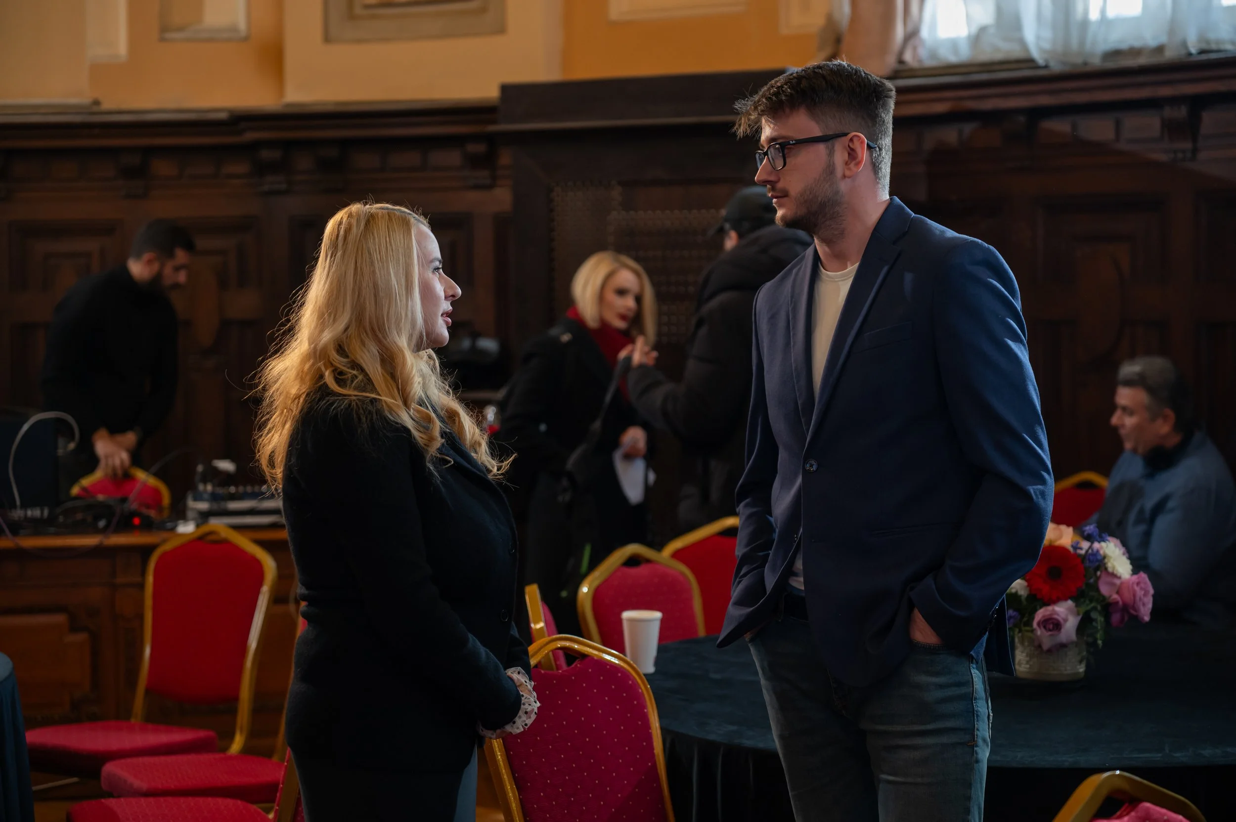 A man and woman are standing face-to-face in a formal setting, possibly a courtroom or conference hall, engaging in a conversation. The woman has long, wavy blonde hair, and the man has short dark hair, glasses, and is wearing a blue blazer. There ar