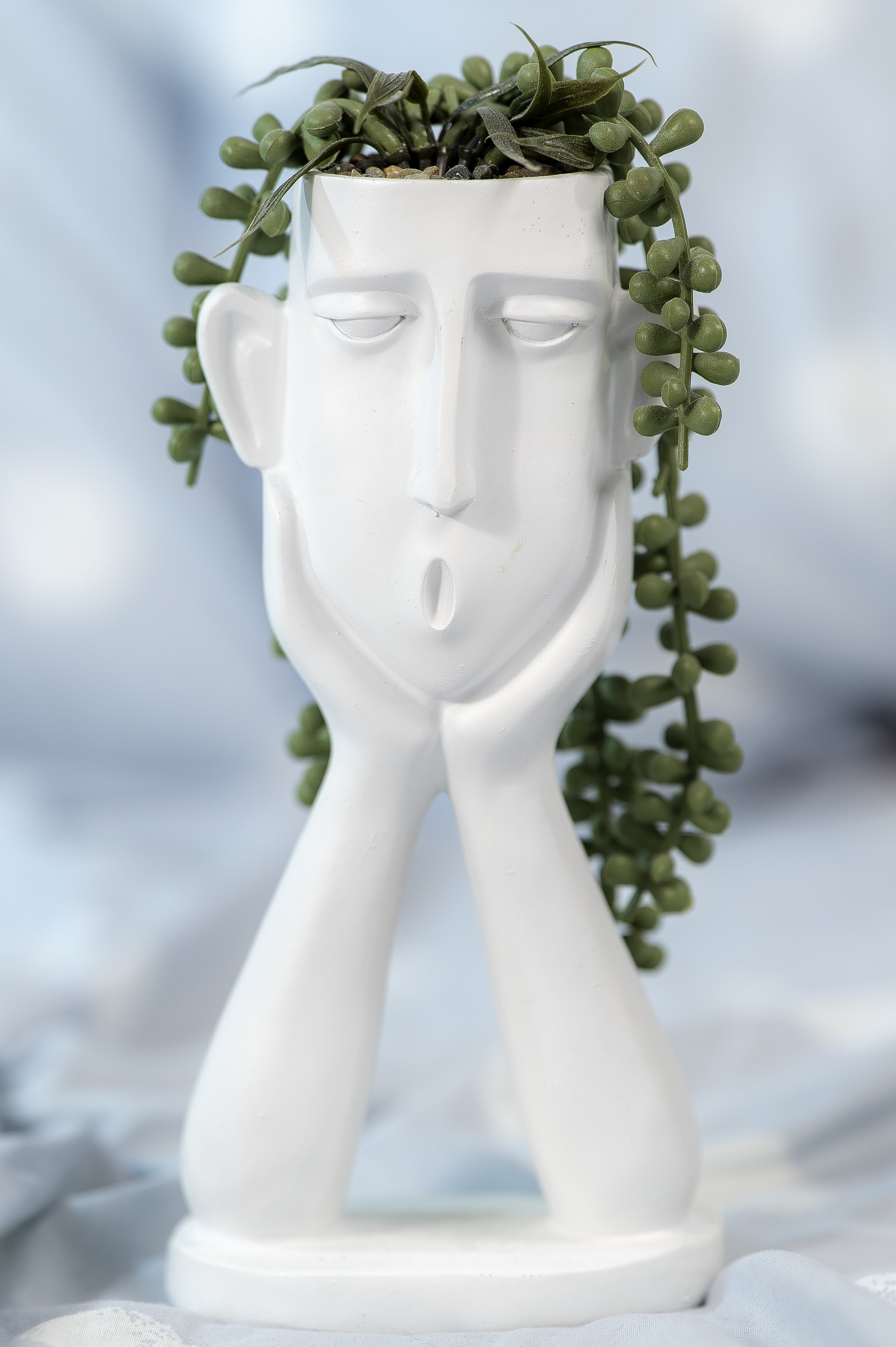 White ceramic figurine of a human face with elongated features, closed eyes, and a slightly open mouth, with trailing green succulent plants on either side and on top, set against a light blue background.
