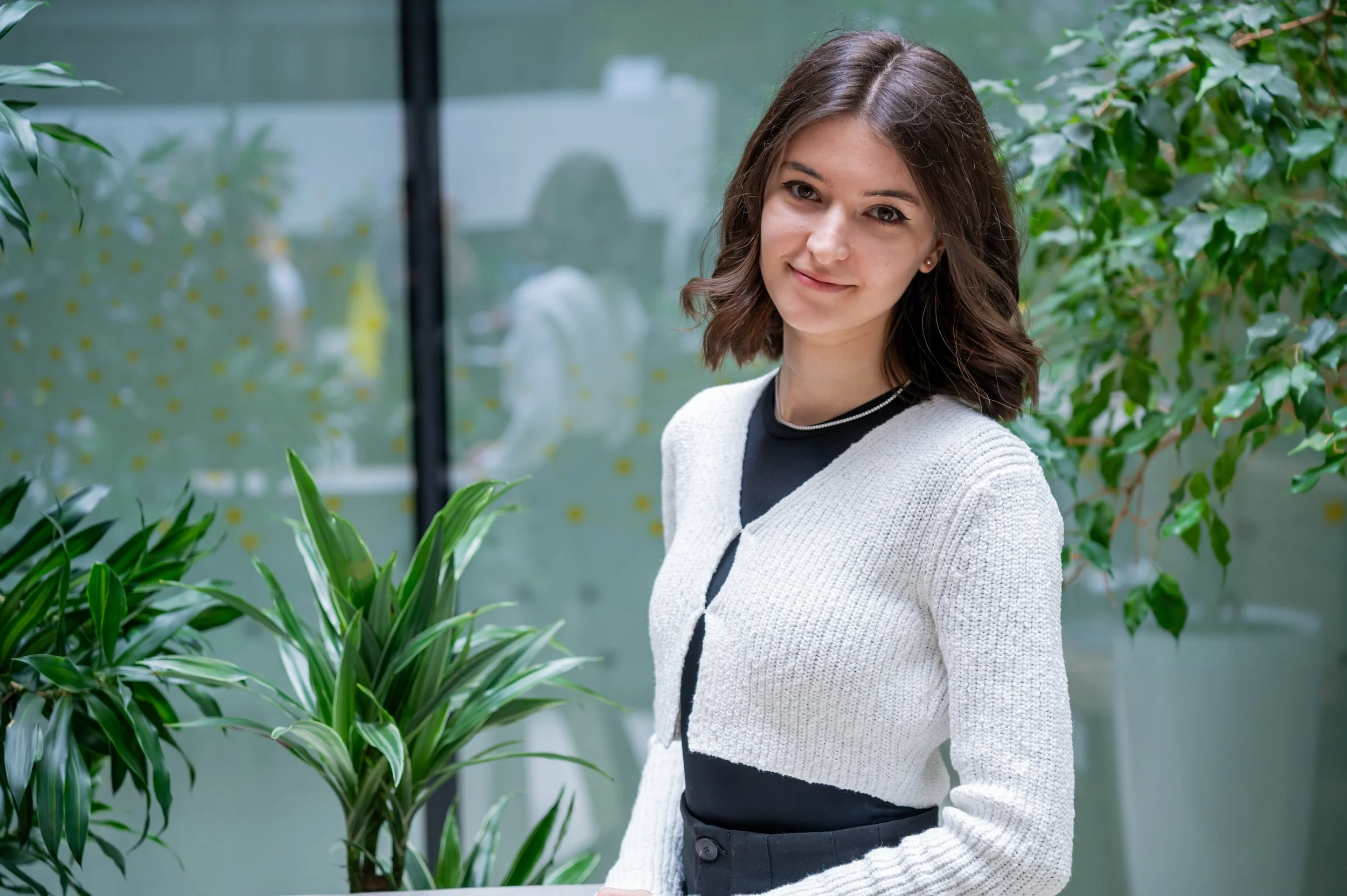 A young woman with shoulder-length brown hair and light skin standing among green plants in a modern indoor setting with a glass wall in the background.