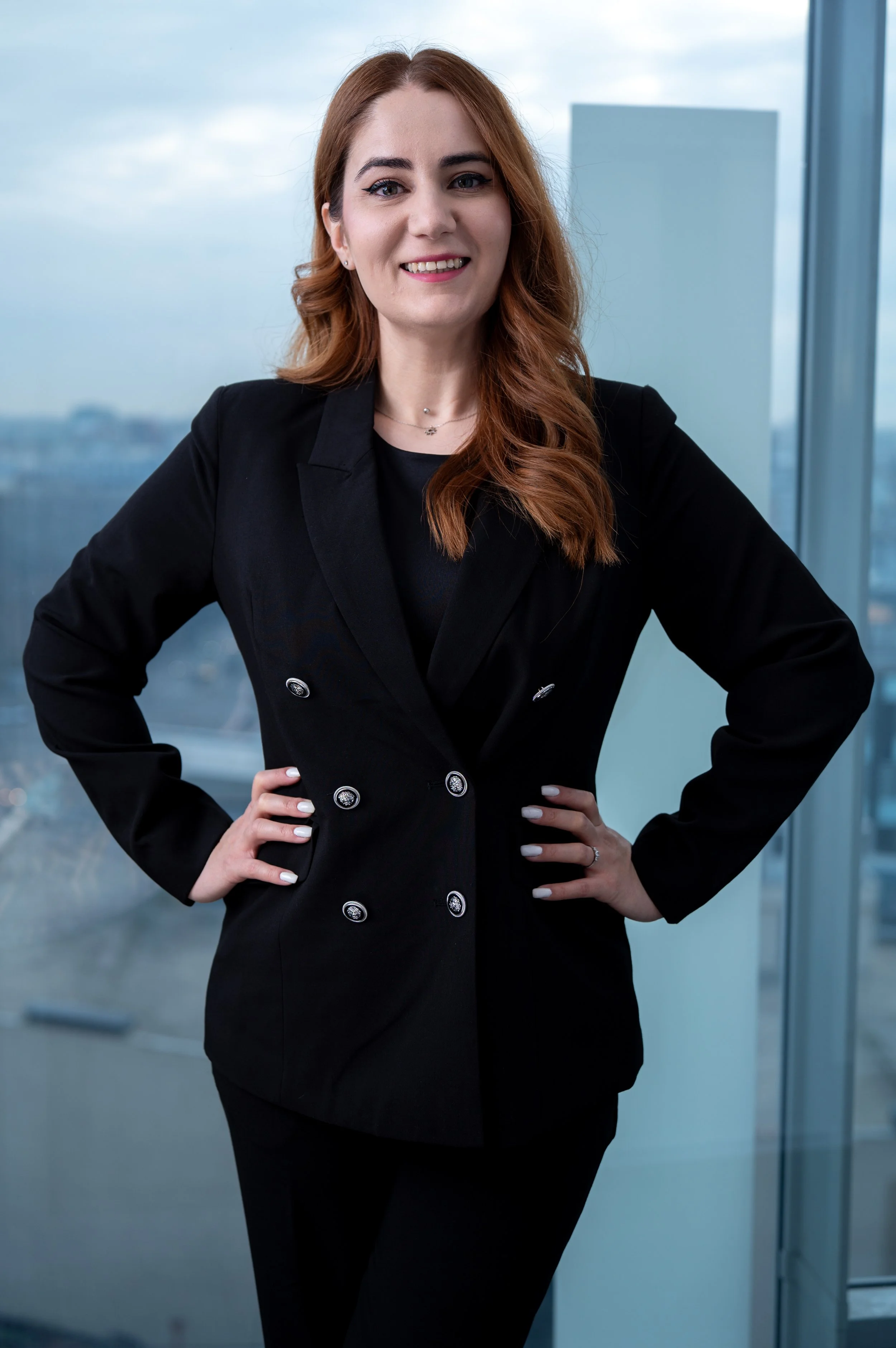 A woman with long, wavy, auburn hair in a black blazer and black pants standing with hands on hips inside a modern office building with large windows showing a cityscape background.