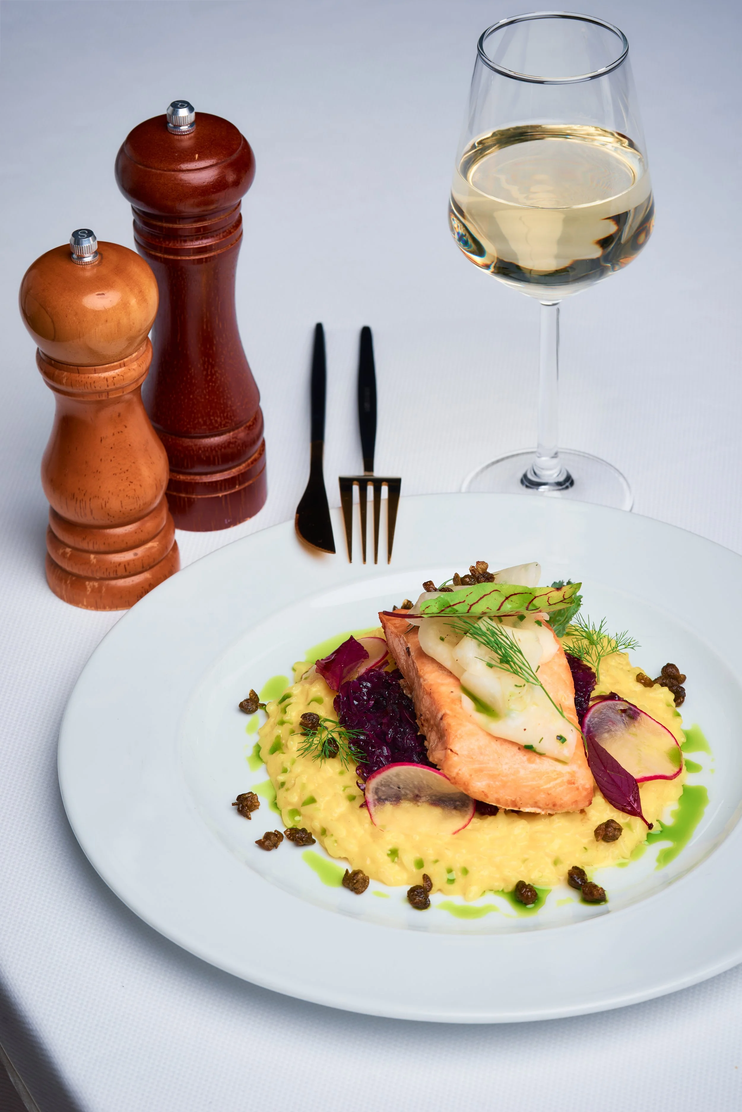 A gourmet meal featuring a piece of cooked salmon topped with garnishes, served on a bed of mashed potatoes with colorful vegetables and sauce. A glass of white wine, salt and pepper shakers, and utensils are on the table.