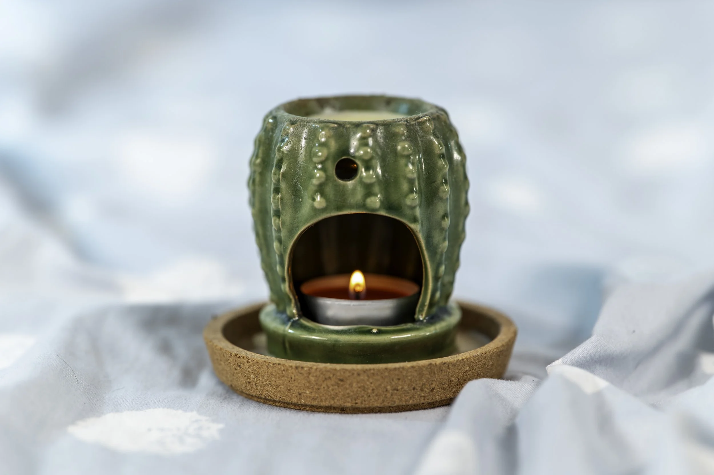Ceramic cactus-shaped candle holder with a lit candle inside, placed on a round cork coaster, against a light blue background with white fabric in the foreground.