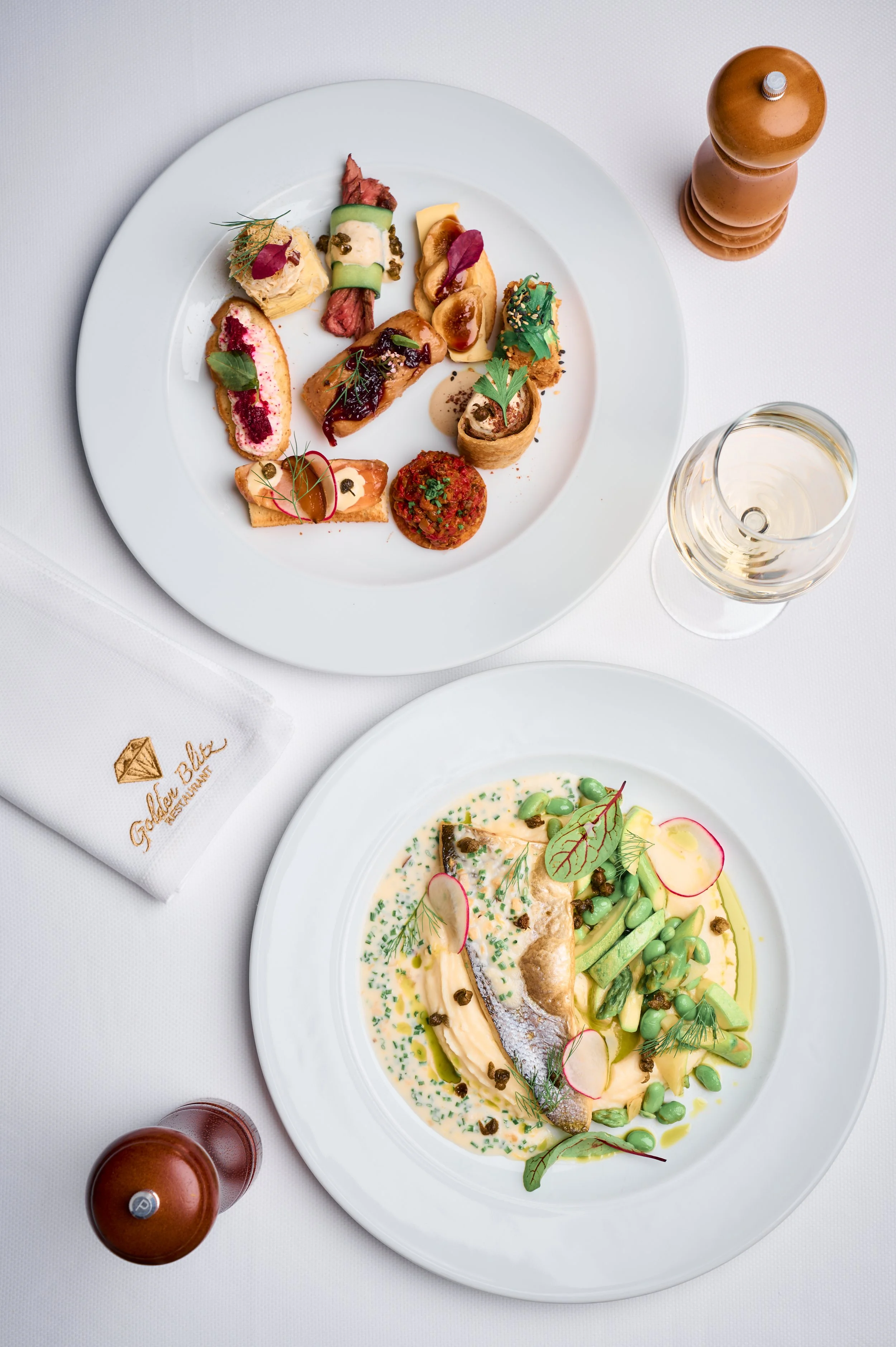 A white plate with various perfectly arranged gourmet appetizers and a glass of white wine.