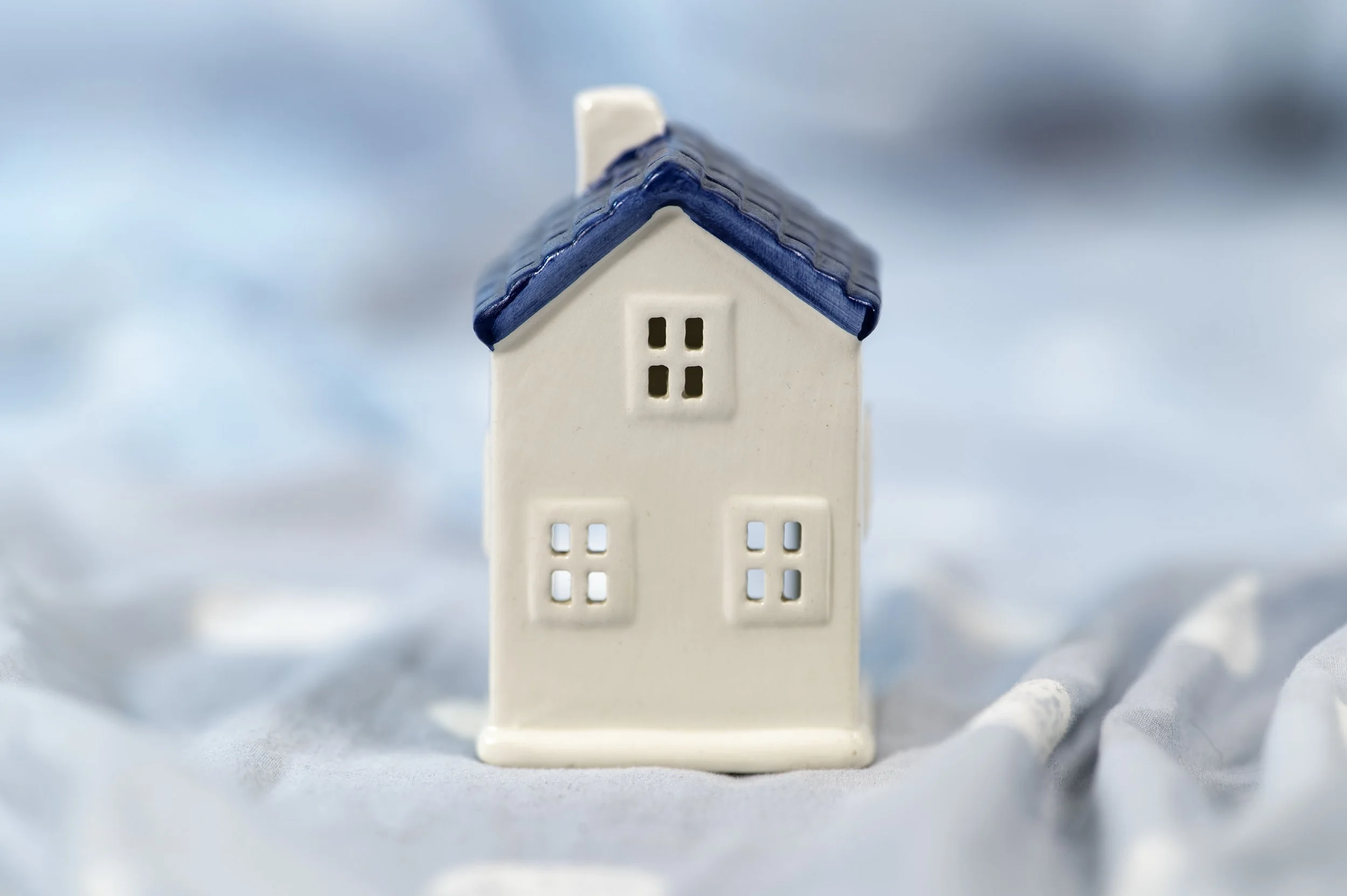 A small decorative ceramic house with a white body and a blue roof, sitting on a soft, cloudy surface.