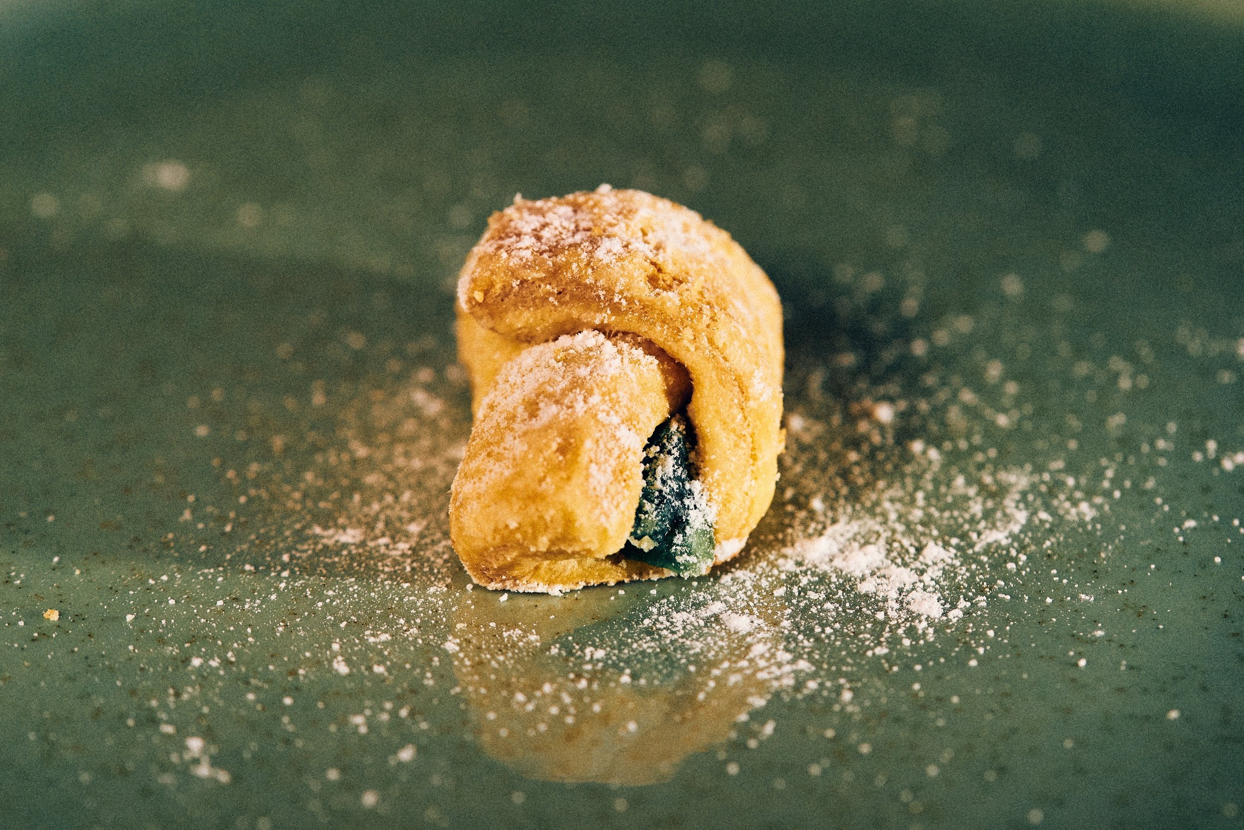 Close-up of a freshly baked cookie with green and black chocolate chips, dusted with powdered sugar, on a green surface with powdered sugar scattered around.