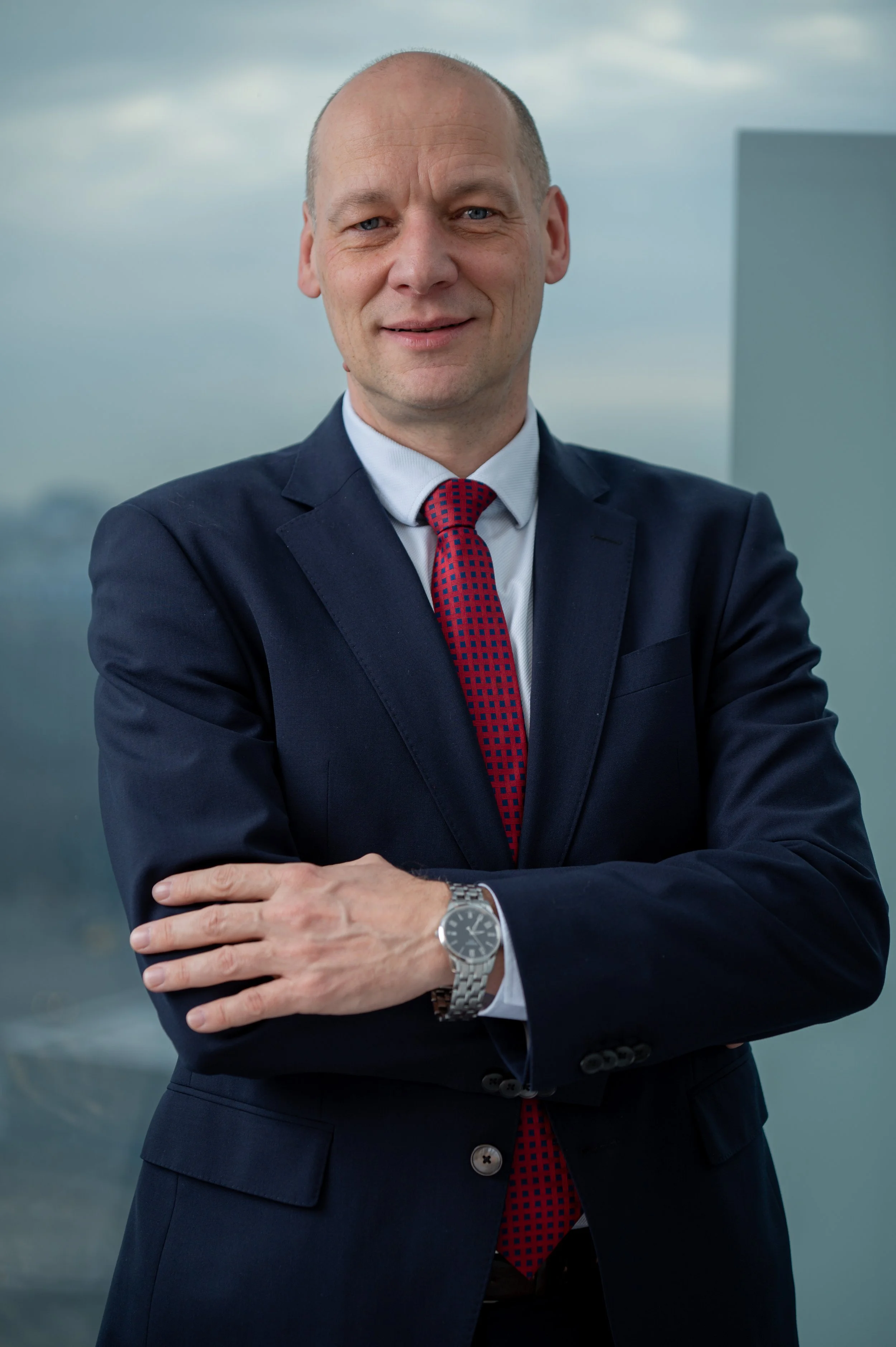 A middle-aged man in a navy business suit, white shirt, and red tie with blue pattern, standing outdoors with his arms crossed, smiling slightly, with a cloudy sky and modern building in the background.