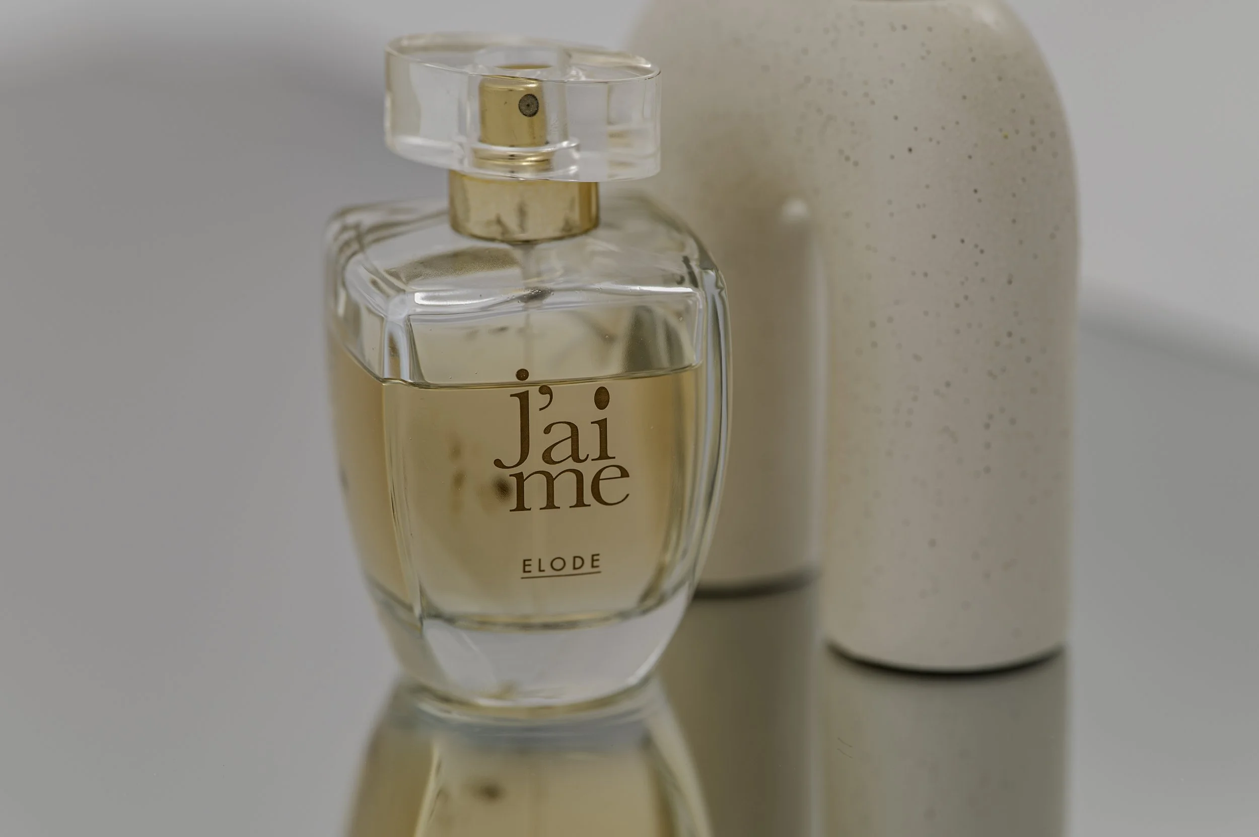 A glass perfume bottle labeled 'J'adore' with a gold and clear cap, placed on a reflective surface, with a speckled beige container in the background.