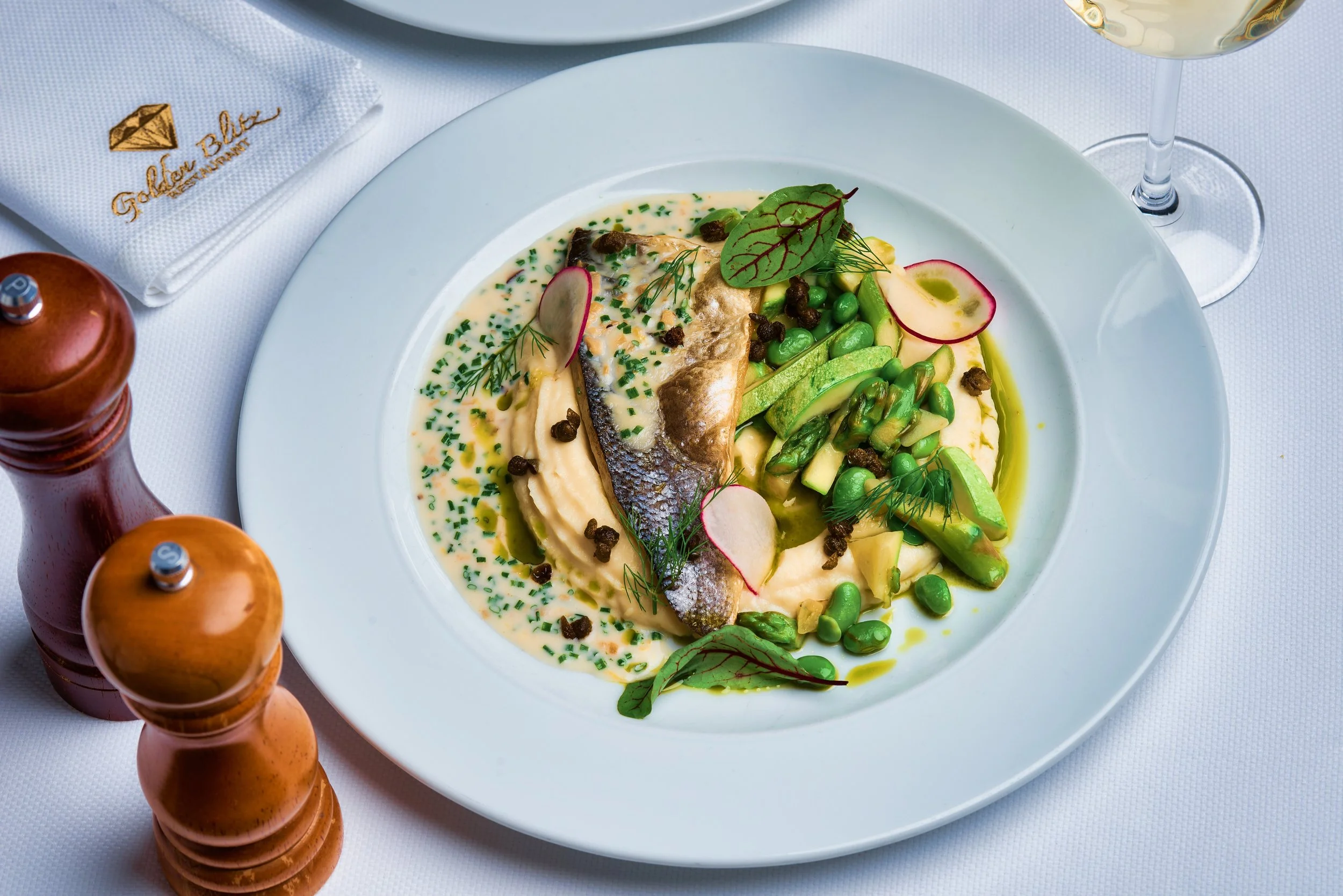 A white plate with fish, mashed potatoes, asparagus, peas, radish slices, and herbs in a creamy sauce, garnished with a leaf. A glass of white wine and salt and pepper shakers are on the table.